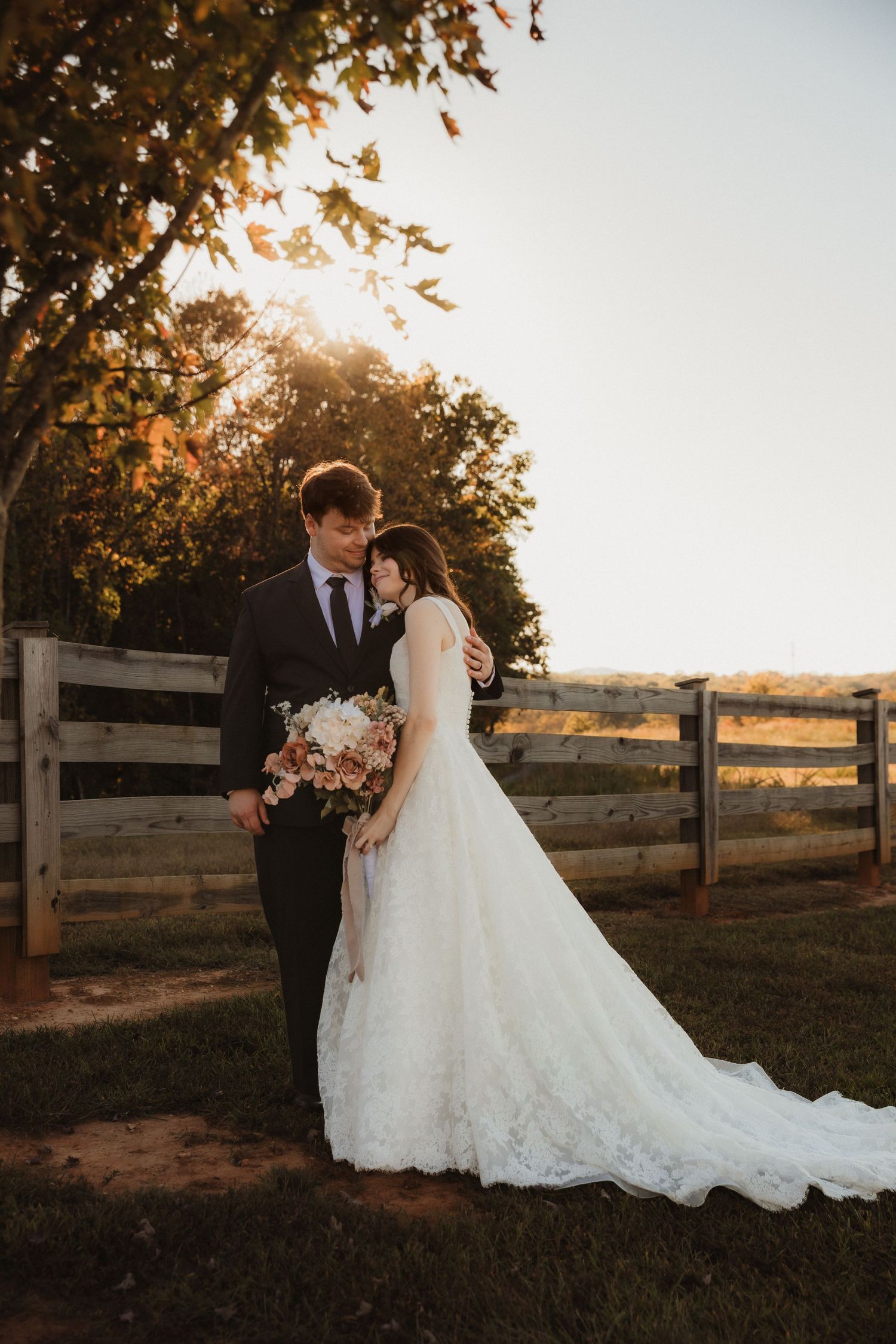 Newlyweds pose against wooden fence during sunset with autumn trees in background.