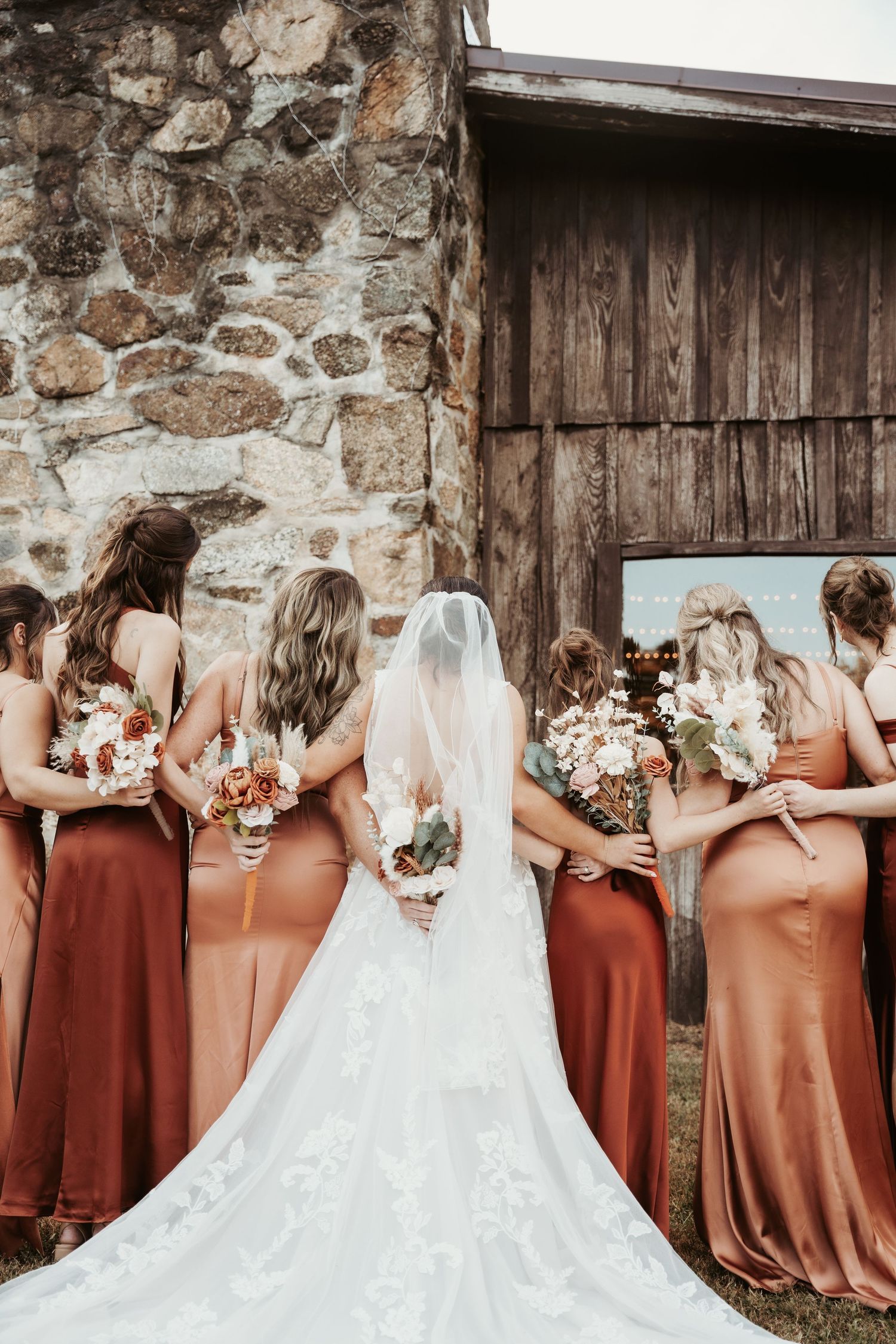 Wedding party in rust-colored dresses posed against rustic stone and wood barn.