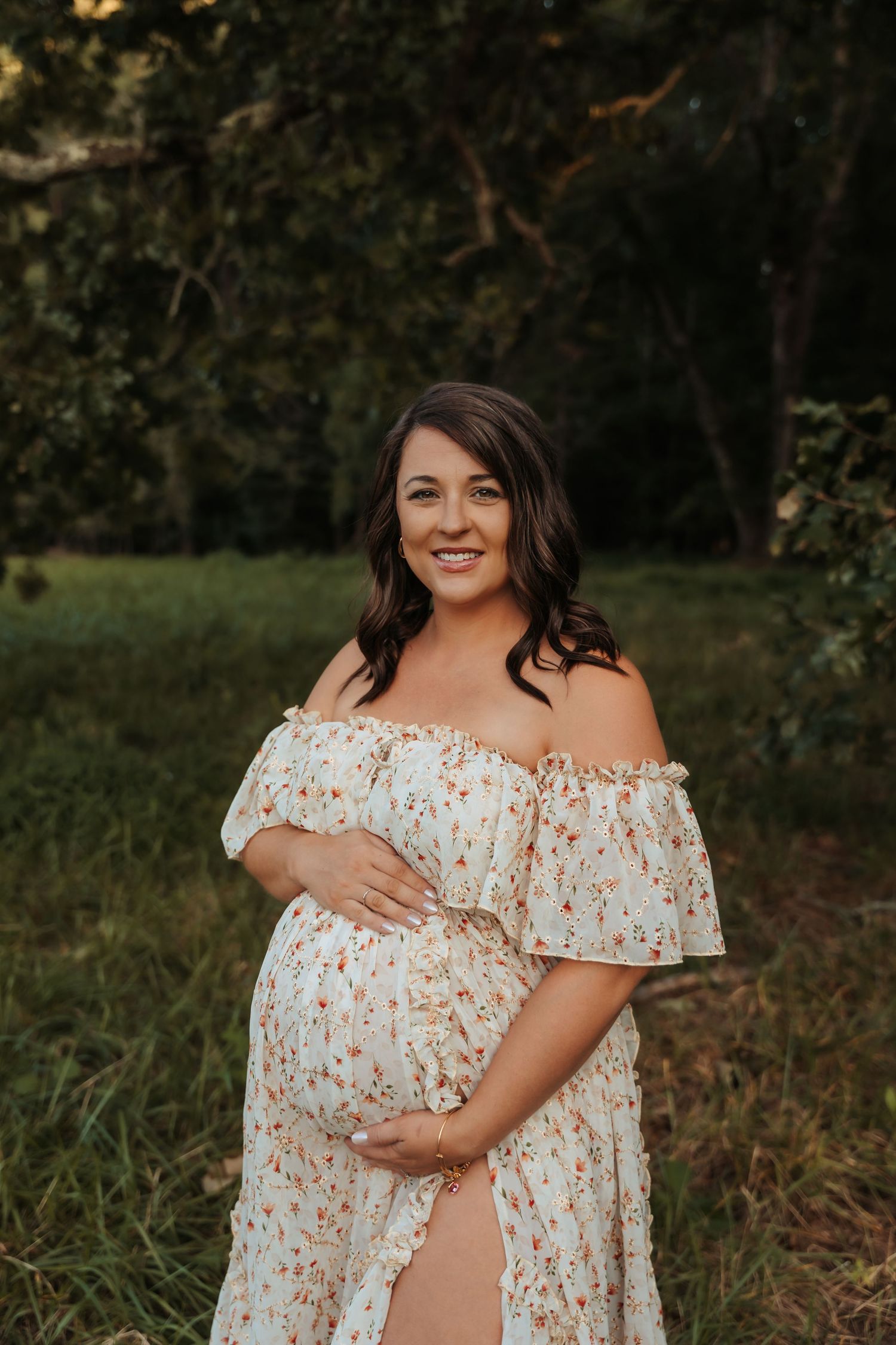 Maternity portrait in a floral off-shoulder dress against a natural outdoor backdrop.