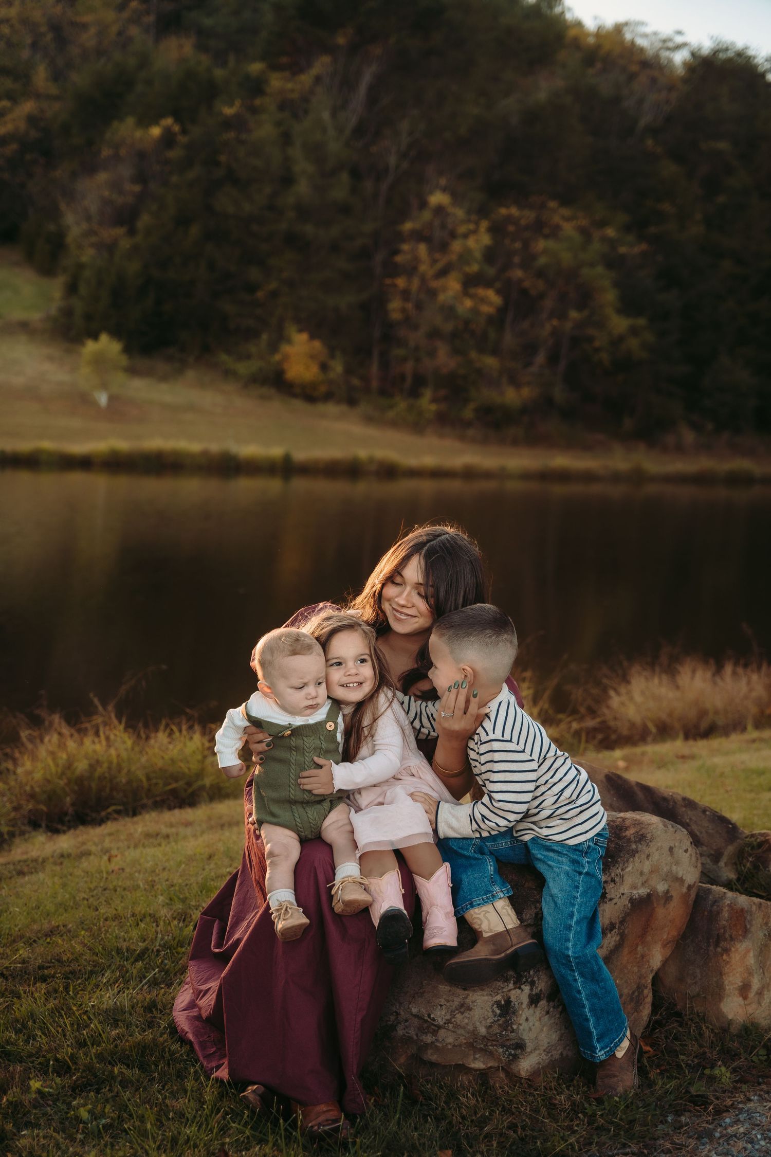 Three children sit together on a large rock by a lake during autumn.