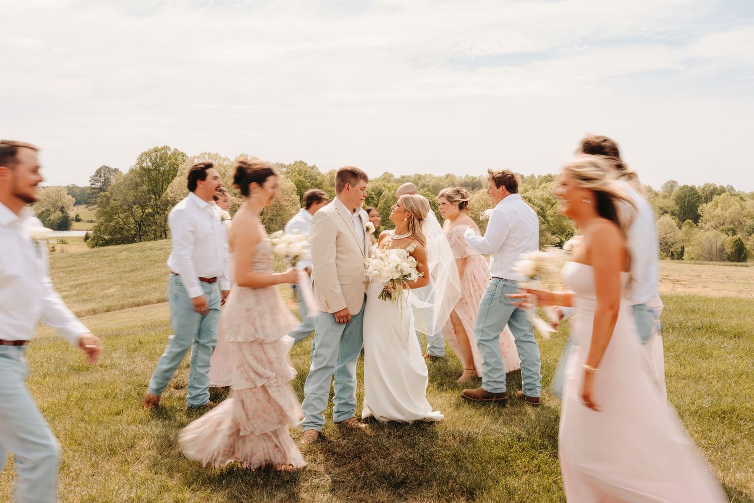 Wedding party walking outdoors on green grass field, bride in white gown with groom and guests in pastel attire.