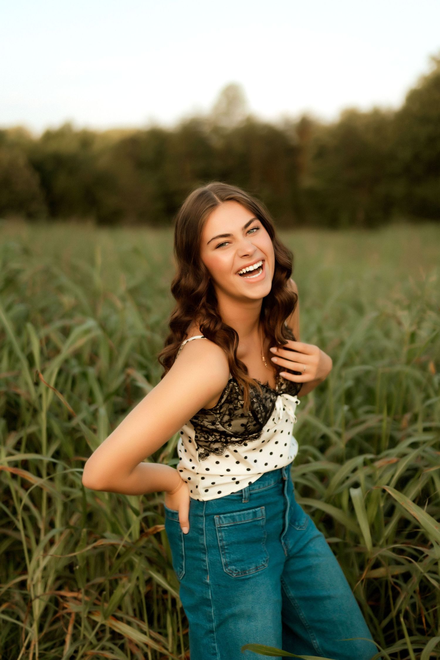Young woman laughing in a green field, wearing polka dot top and blue jeans, golden hour outdoor portrait.
