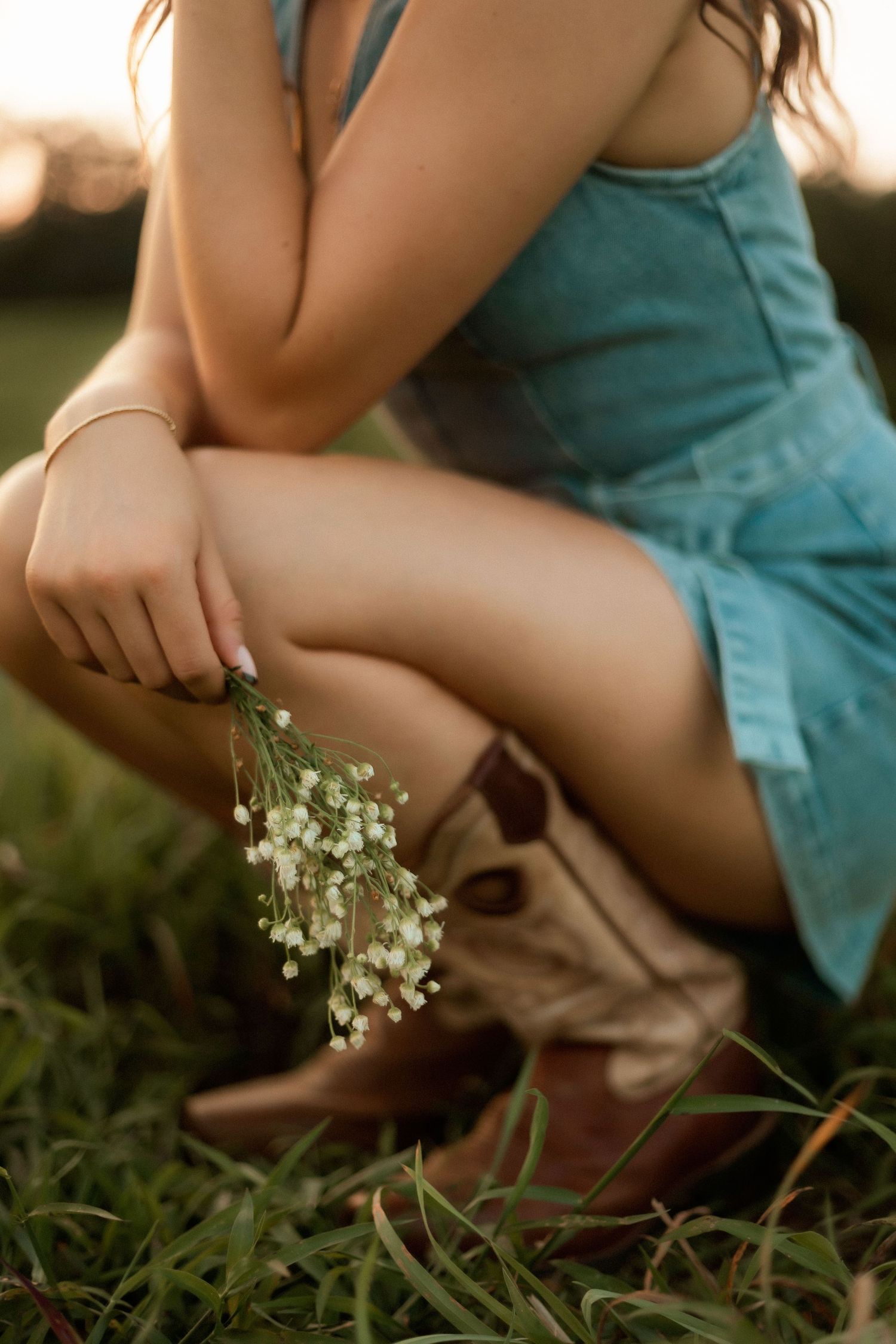 Woman in teal dress and cowboy boots crouching in grass holding small white wildflowers at golden hour.