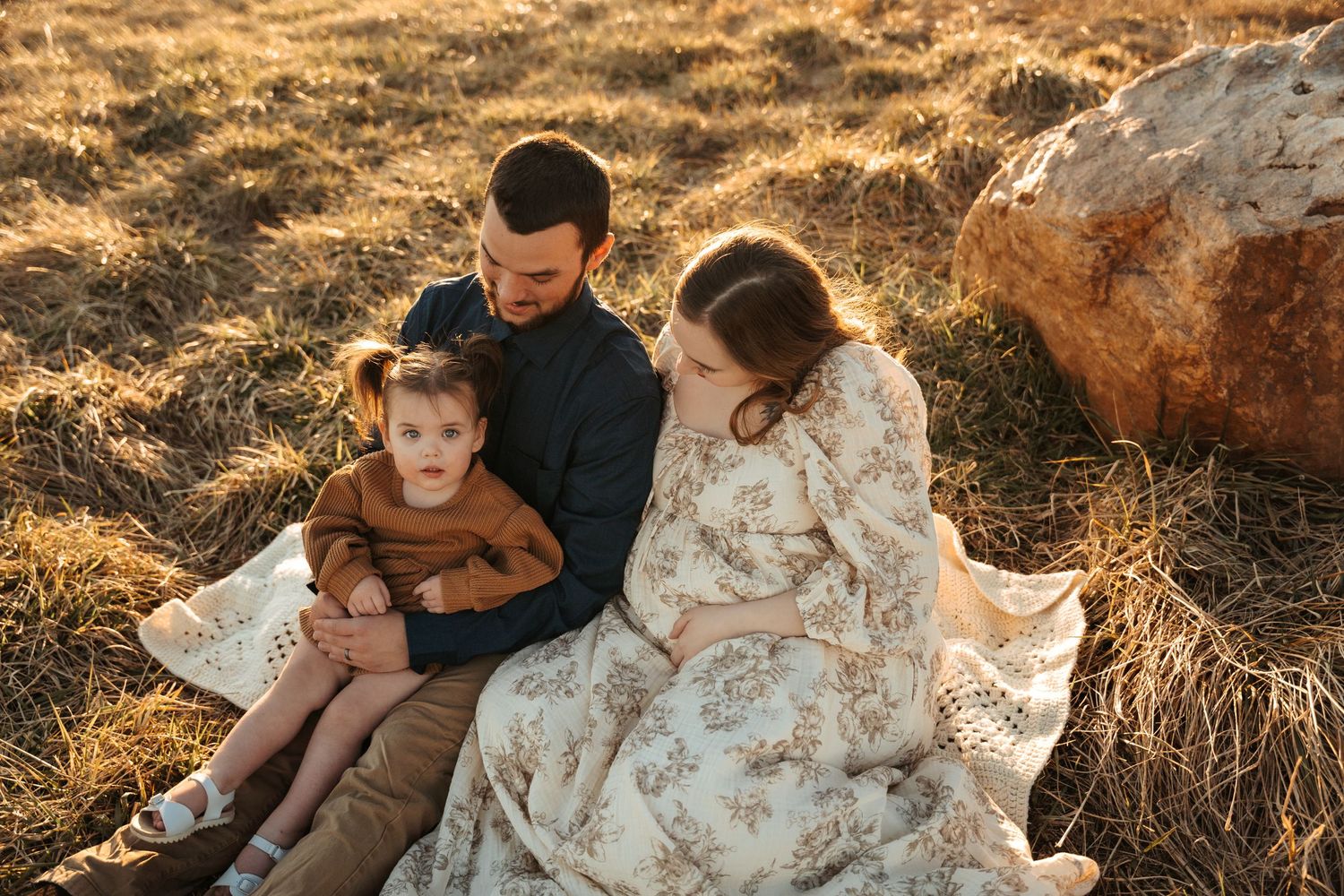 Family sits together on a blanket in a golden field during sunset.