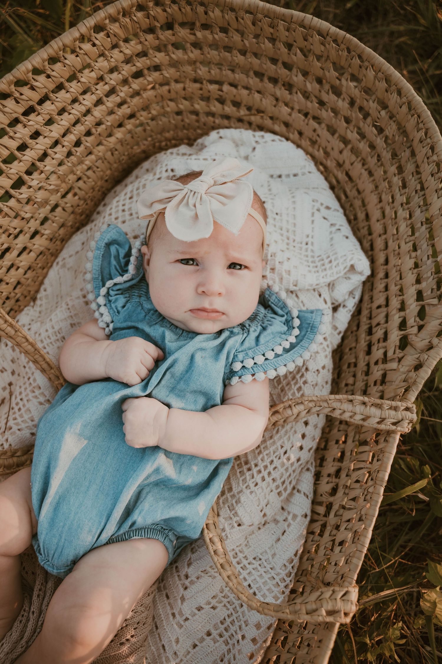 Baby in blue romper resting in woven basket outdoors.
