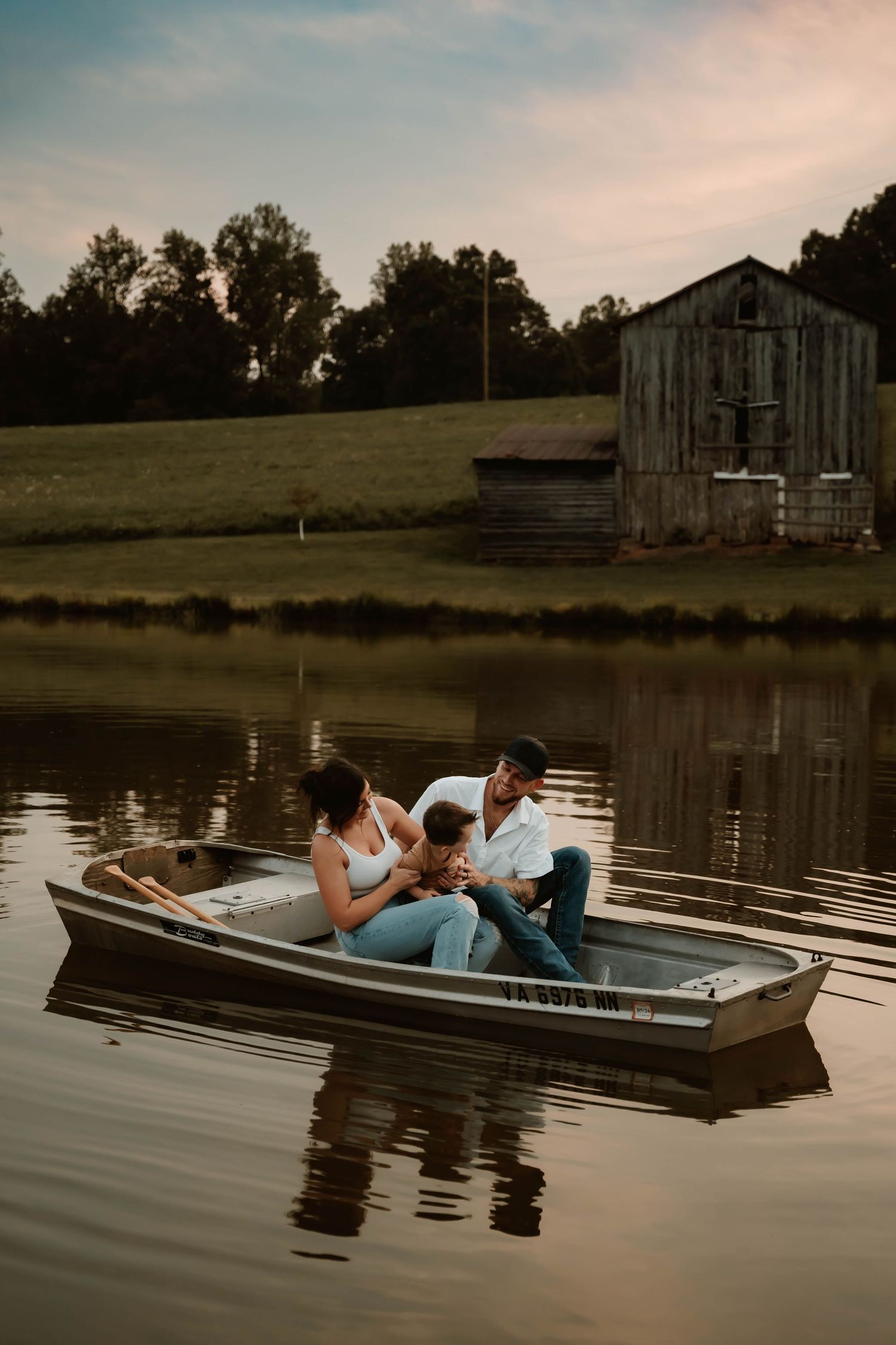 Romantic couple lying in wooden boat on calm lake at sunset near rustic barn.