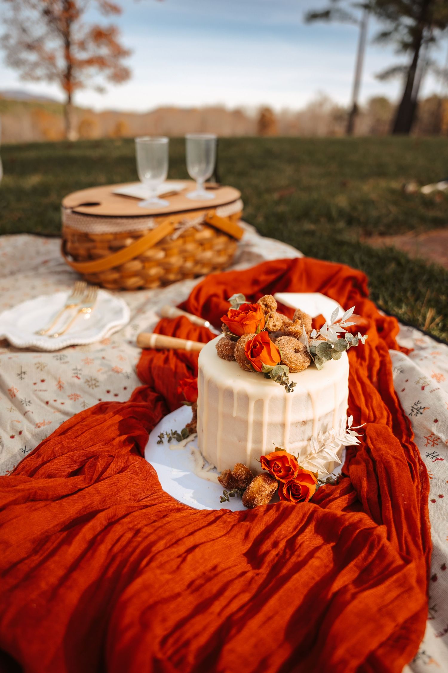 A white cake decorated with dried flowers on an orange fabric with picnic setting.