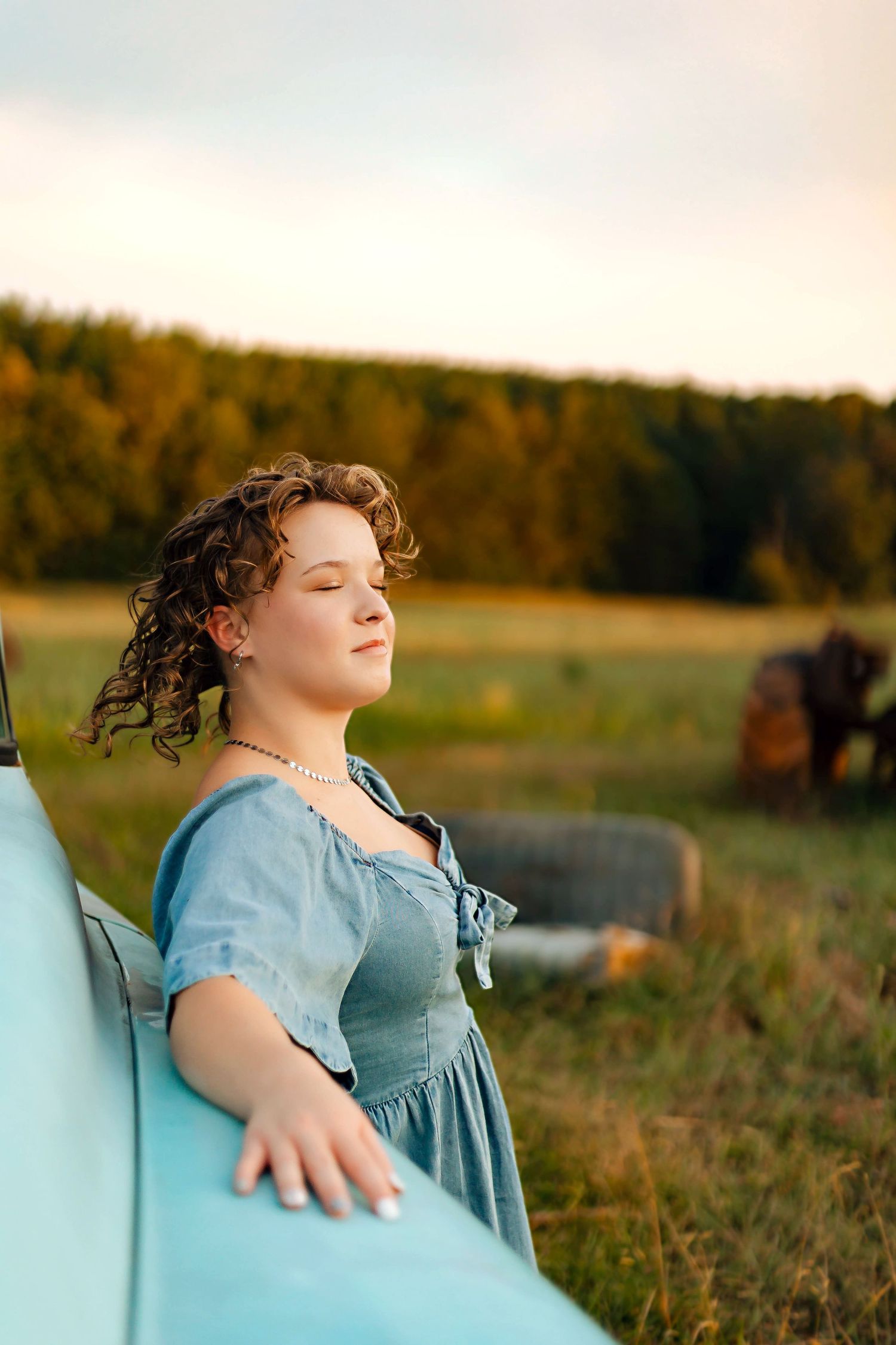 Young woman in blue dress leaning on vintage turquoise truck in golden hour field with hay bales.