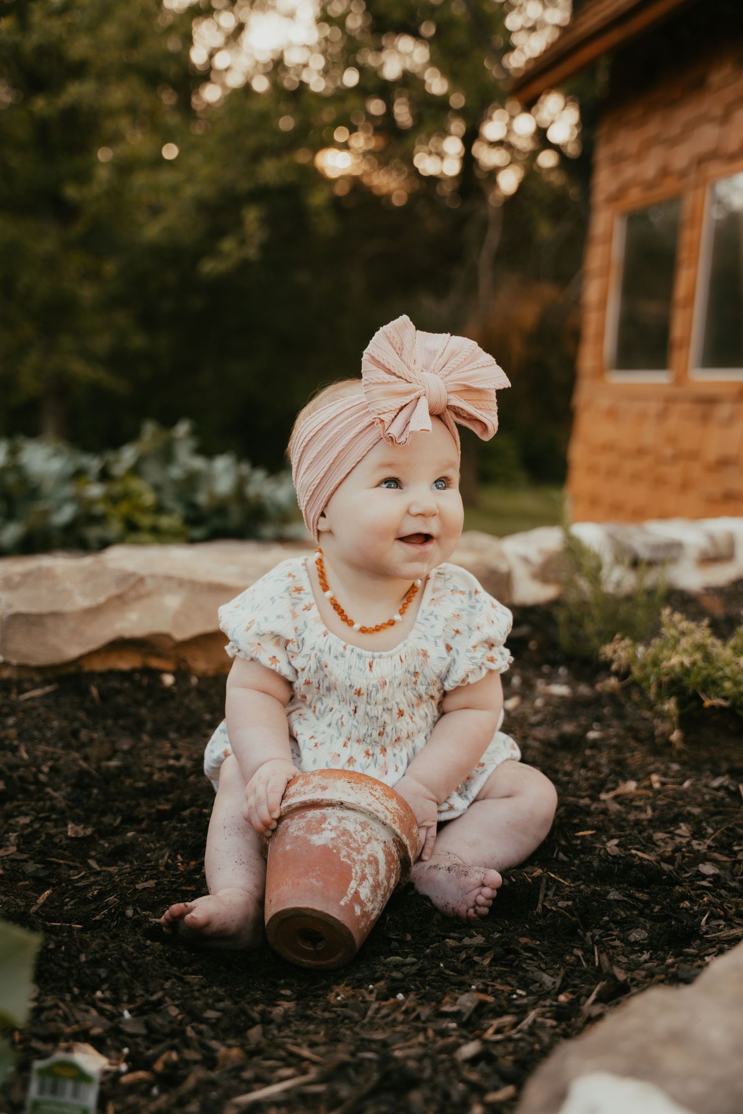 Baby wearing pink bow headband sitting in garden mulch near stone wall.