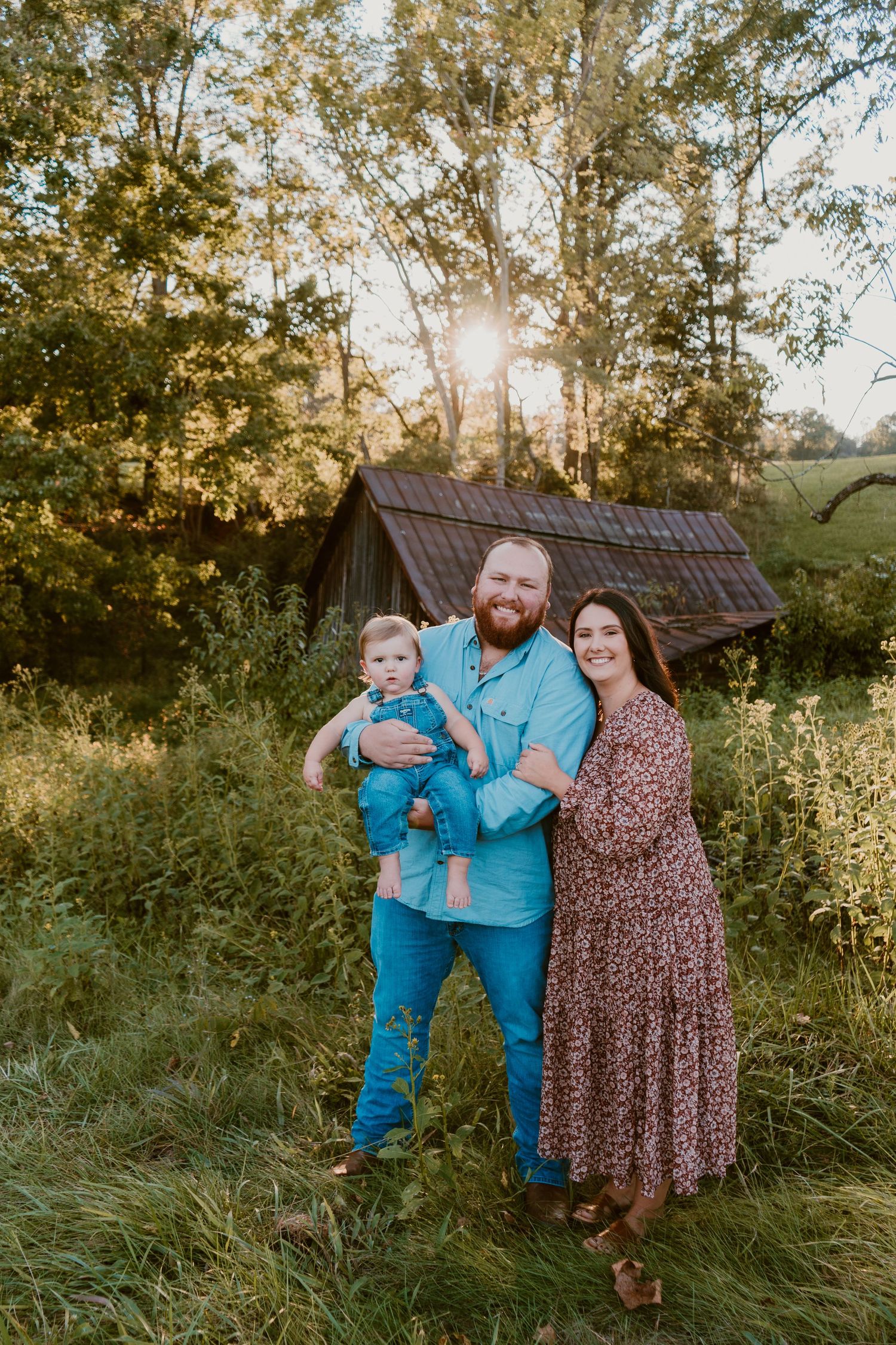 Family portrait in casual attire against a rustic barn backdrop during golden hour.