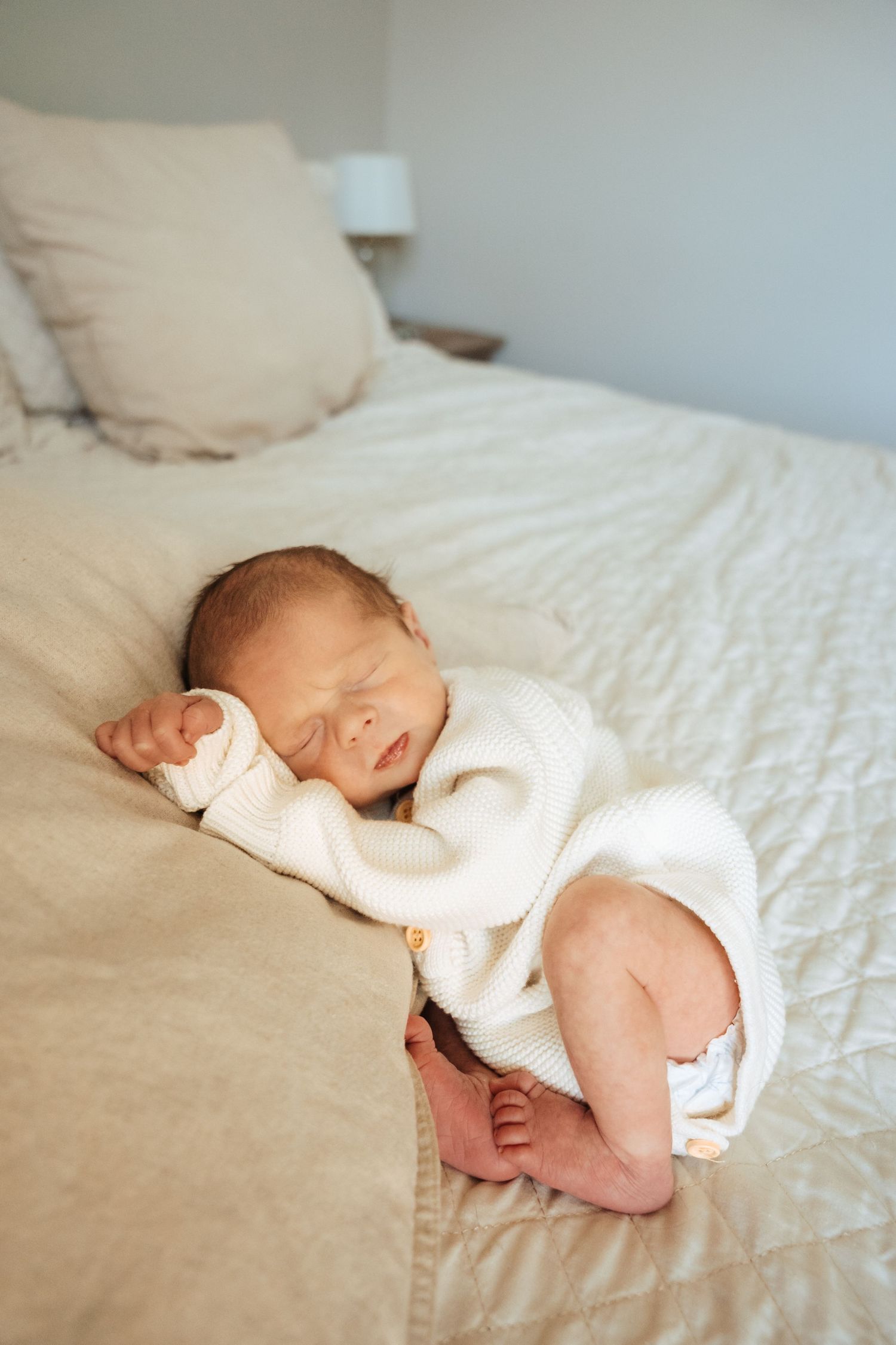 Sleeping newborn baby wearing white outfit rests peacefully on white bedding.