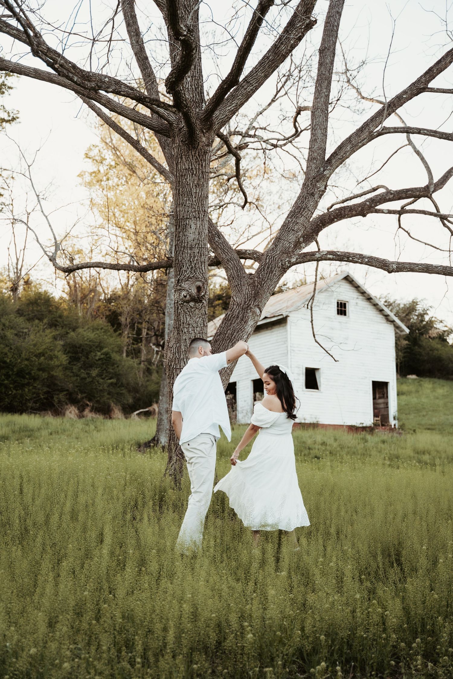 A couple dances in a field near a white farmhouse at golden hour.