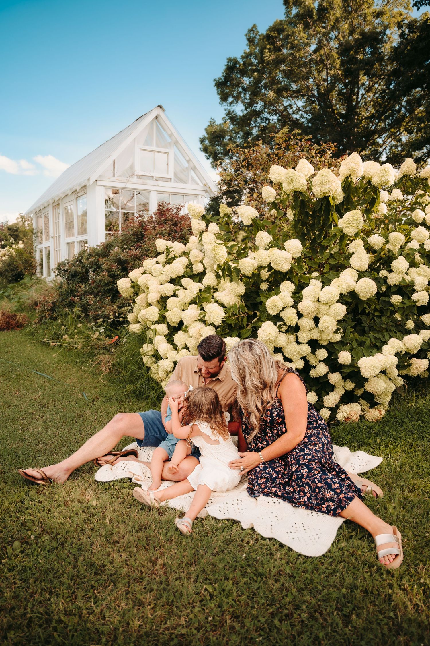 Family relaxing on blanket by greenhouse surrounded by blooming white hydrangeas.