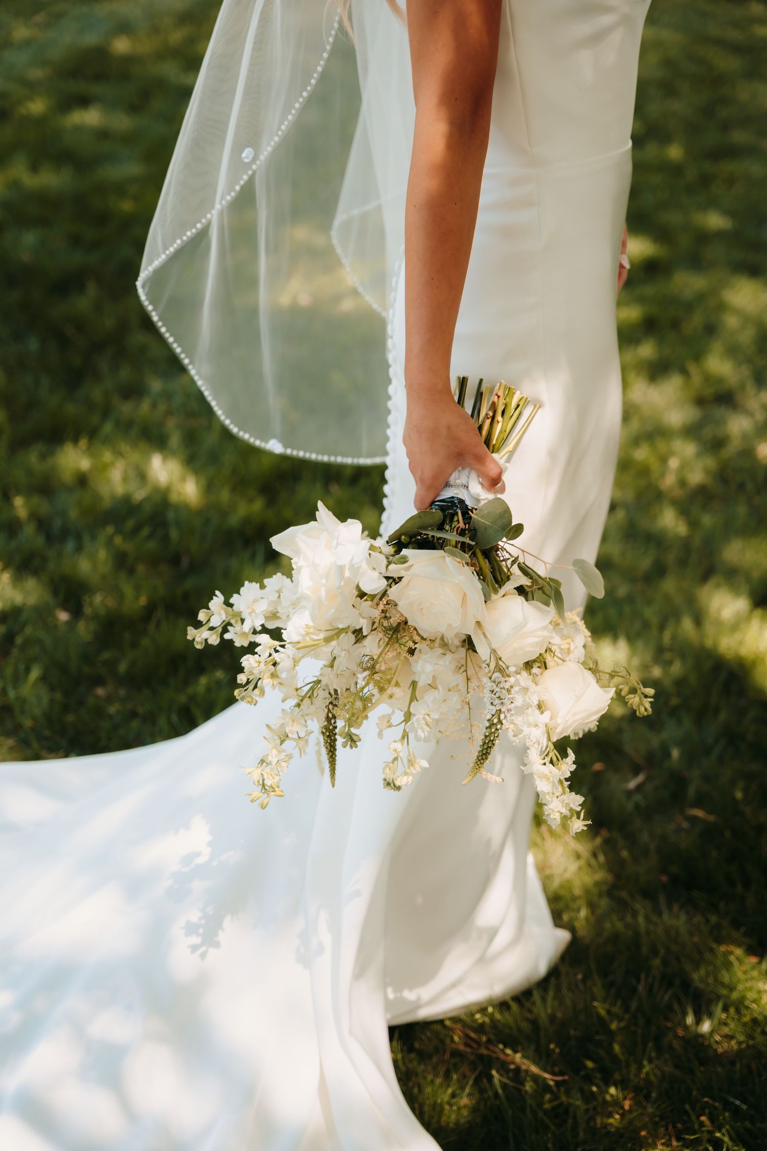 Bride in white gown holding elegant white peony bouquet with greenery, veil flowing on sunlit grass.