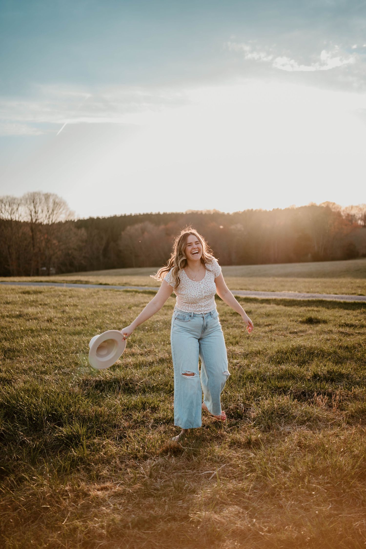 Person in casual attire dancing with a hat in a golden field during sunset.