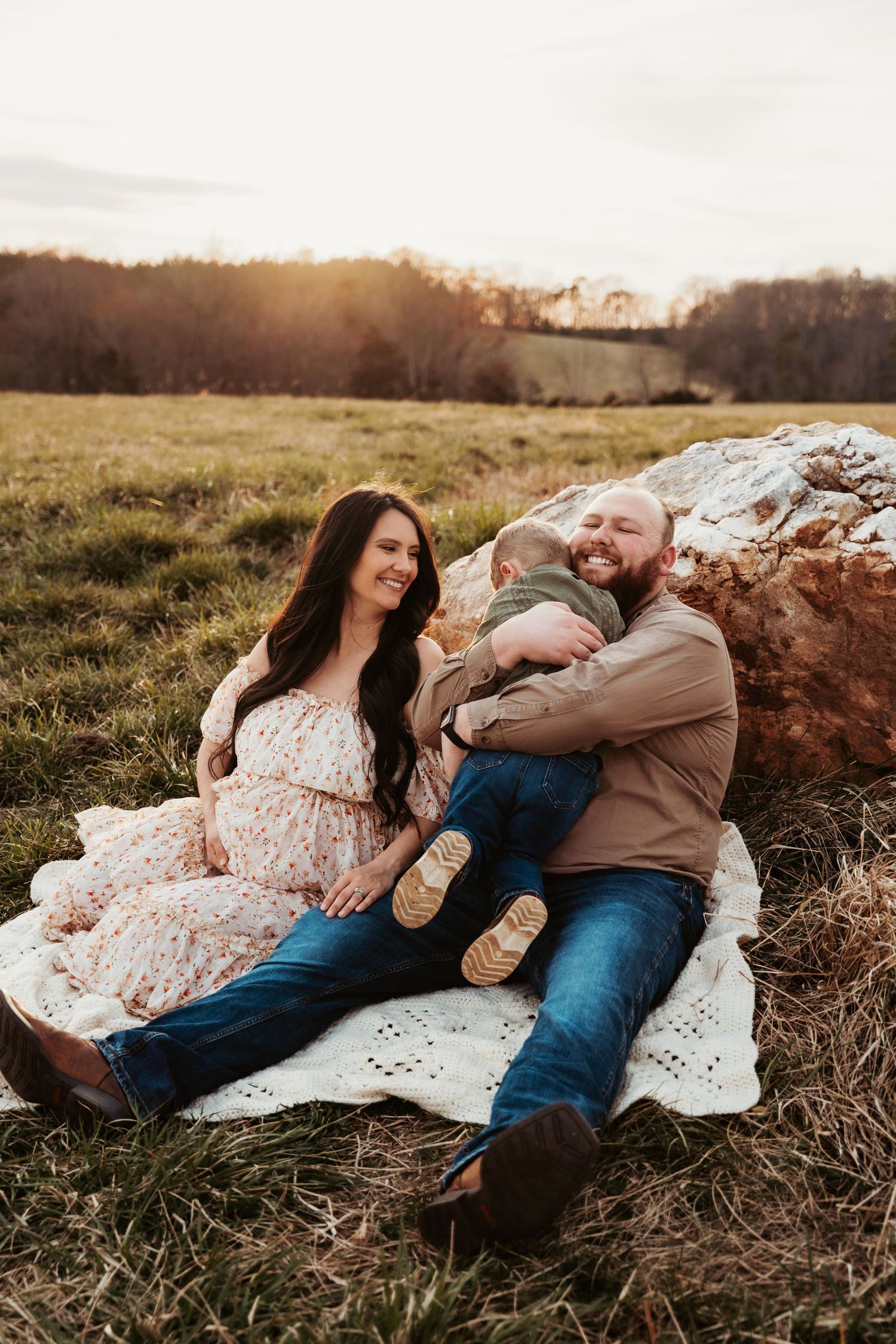 Family relaxing on blanket by large rock in field during golden hour.