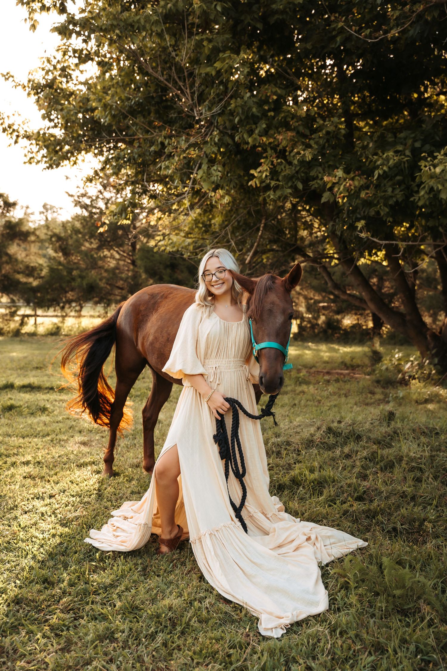 Woman in cream dress stands with brown horse in golden hour sunlight among green trees.