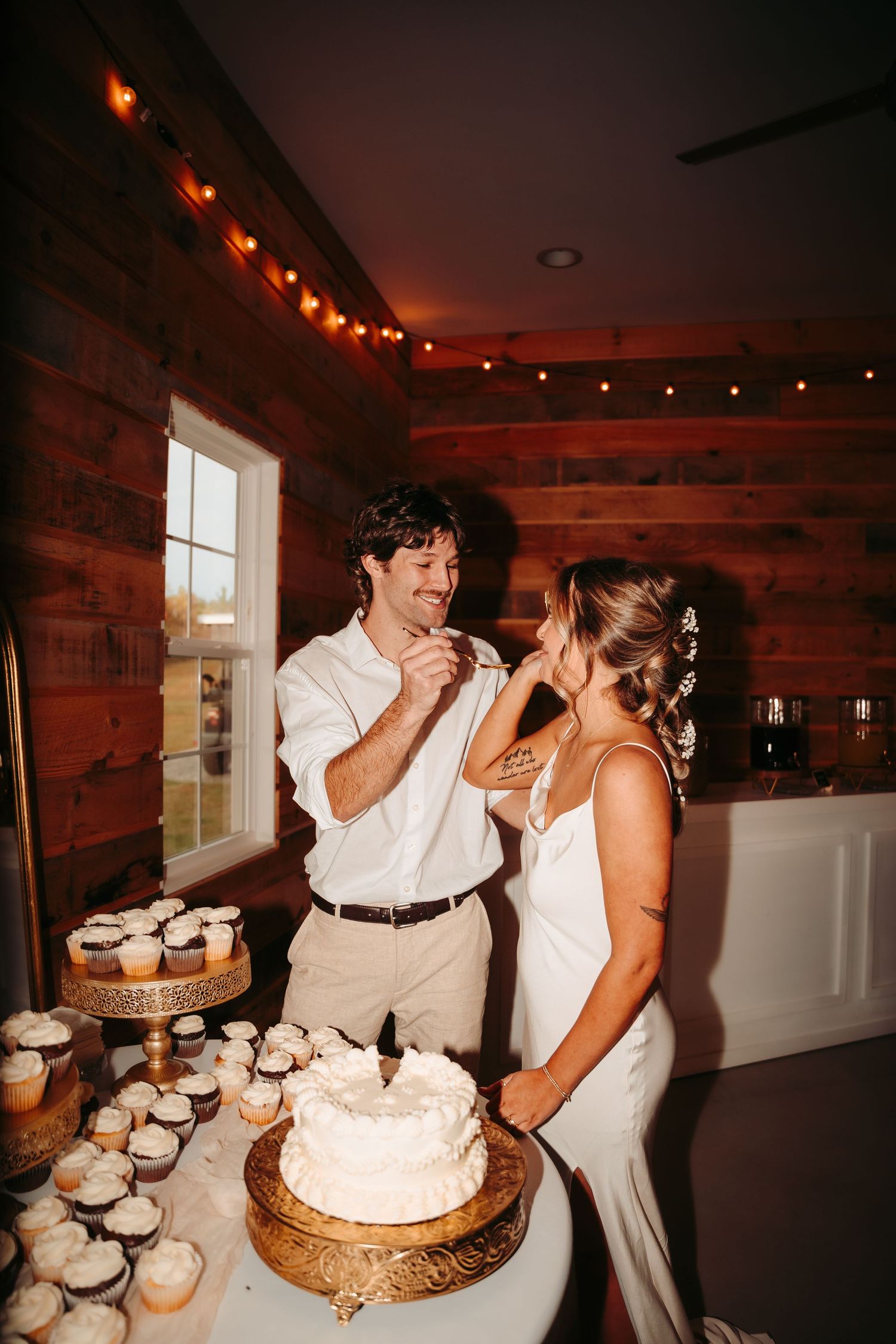 Wedding cake cutting and feeding moment in rustic venue with string lights.