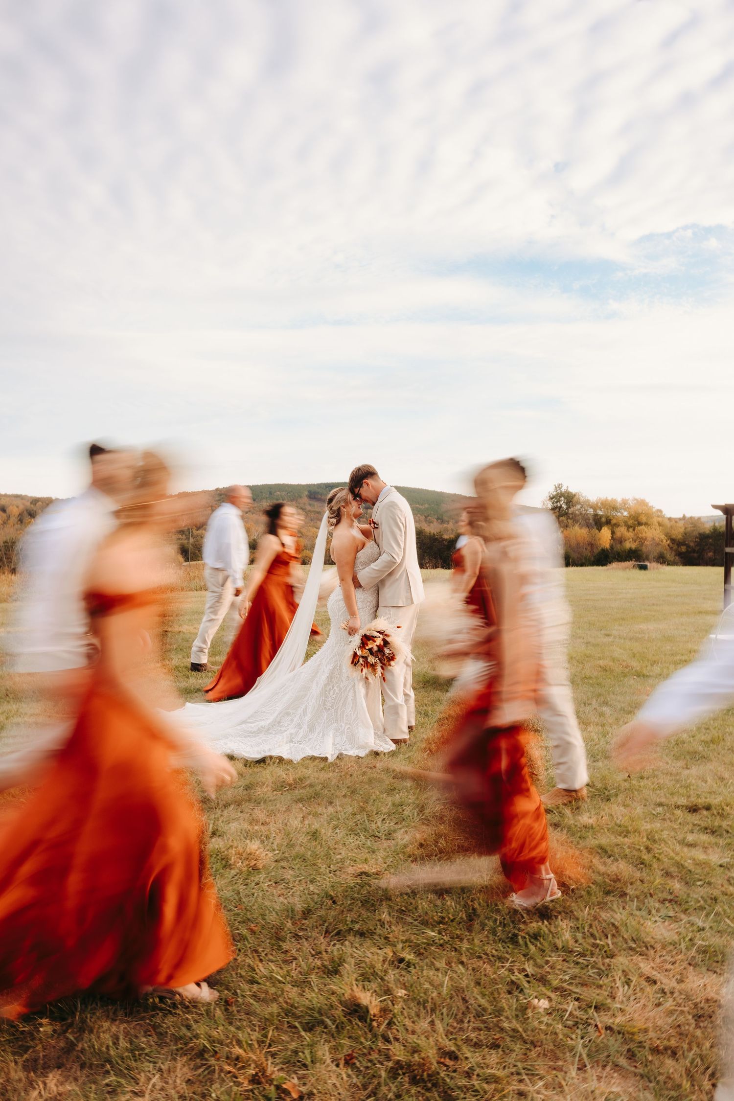 Bride and groom share a kiss outdoors while blurred wedding party in rust orange dresses moves around them on a green field.