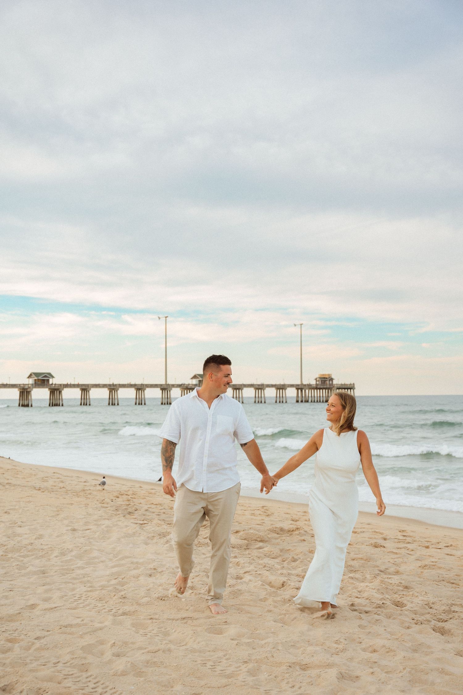 Couple in white outfits holding hands and walking on a sandy beach with a pier in the background.