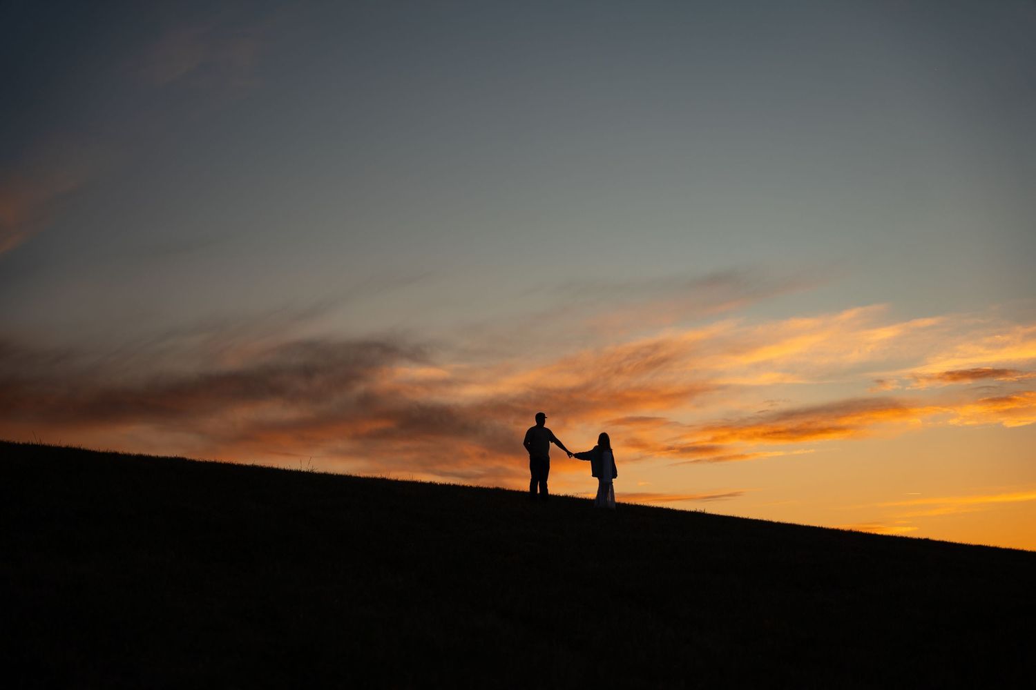 Silhouette of couple holding hands on a hill at sunset with vibrant orange and blue sky backdrop.