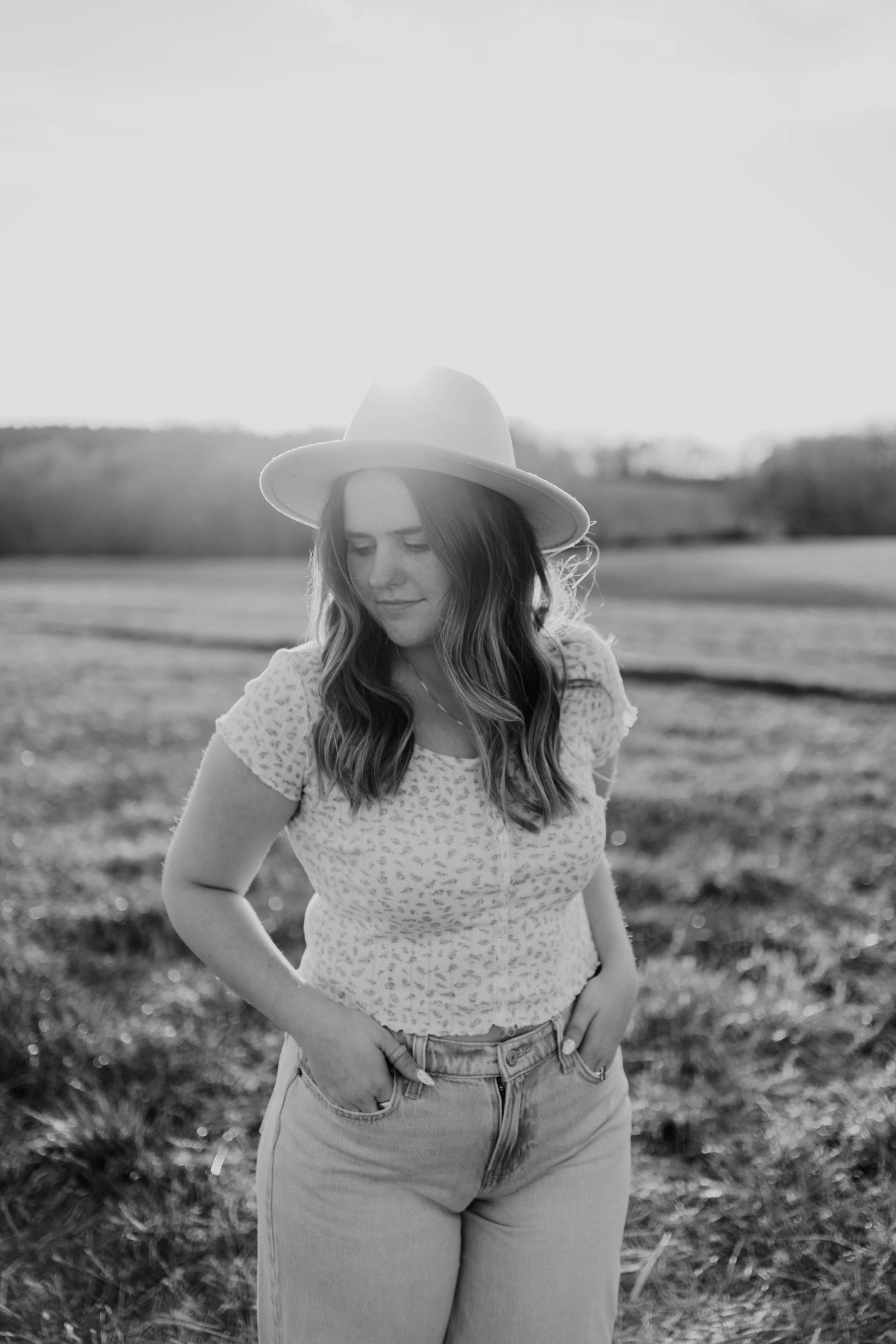 Black and white portrait of someone wearing a hat and floral top in a rural setting.
