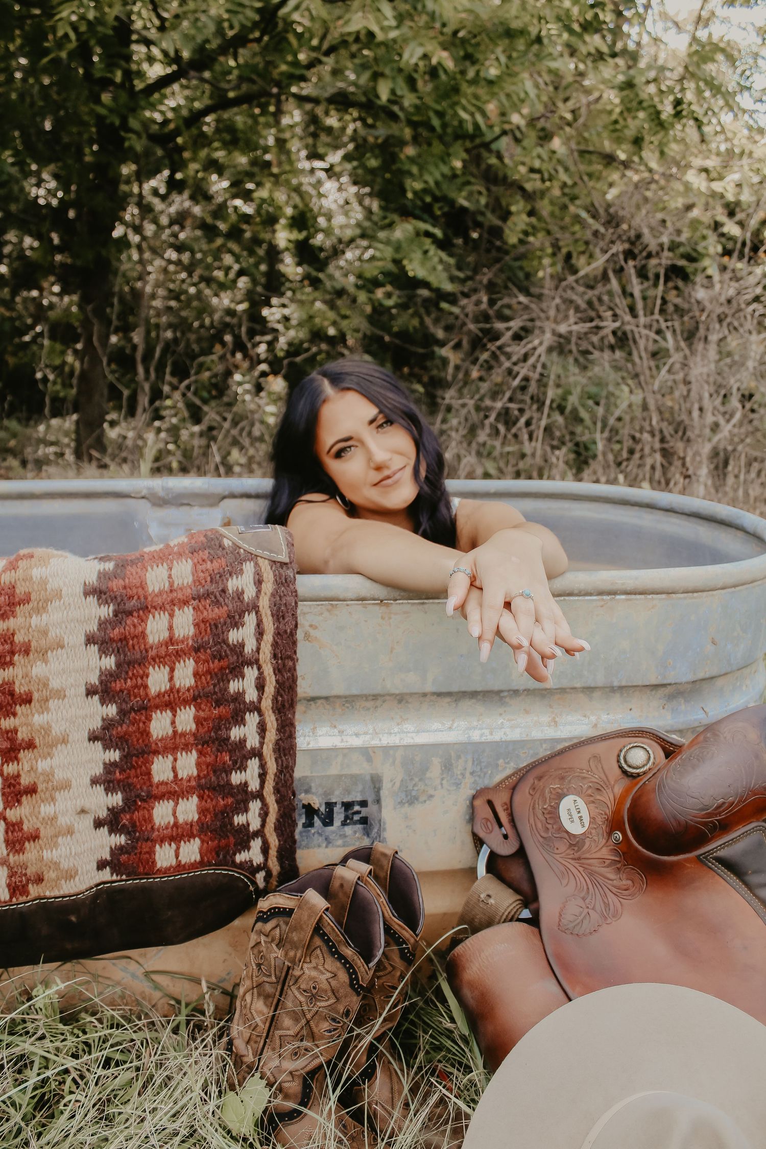Western-themed portrait with stock tank, saddle, and southwestern blanket in outdoor setting.