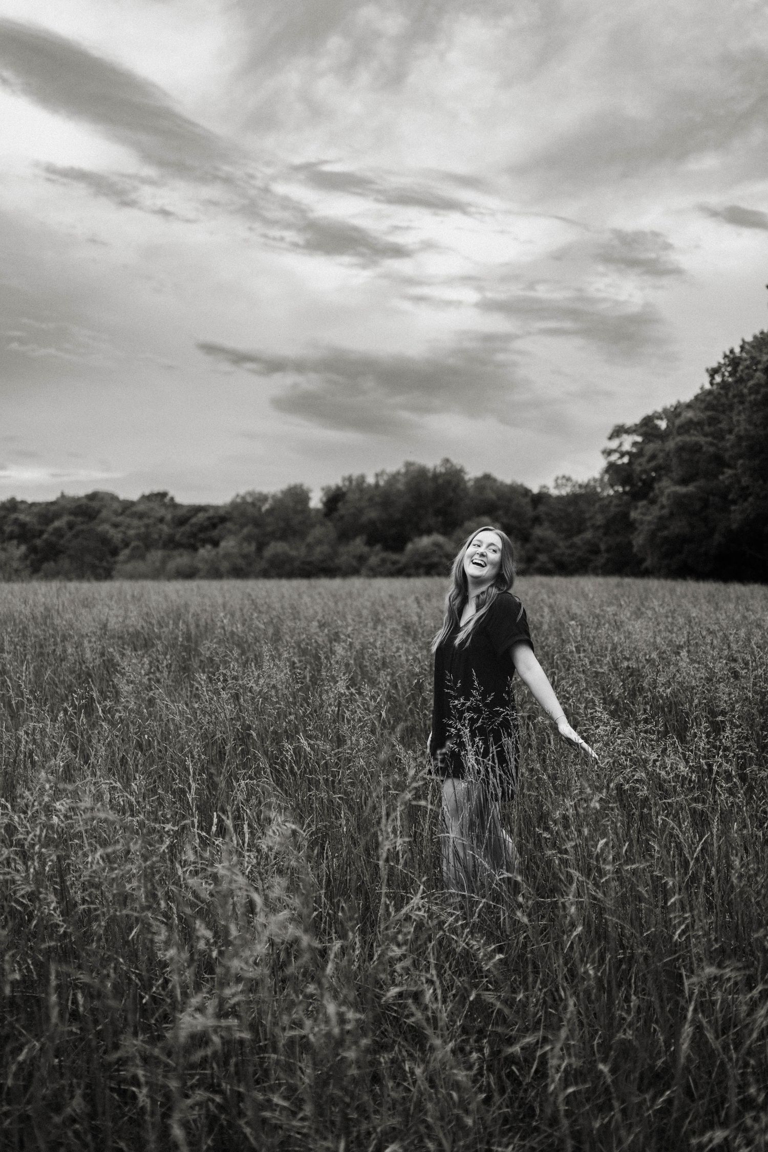 Black and white portrait of someone joyfully dancing in a field with dramatic cloudy sky overhead.