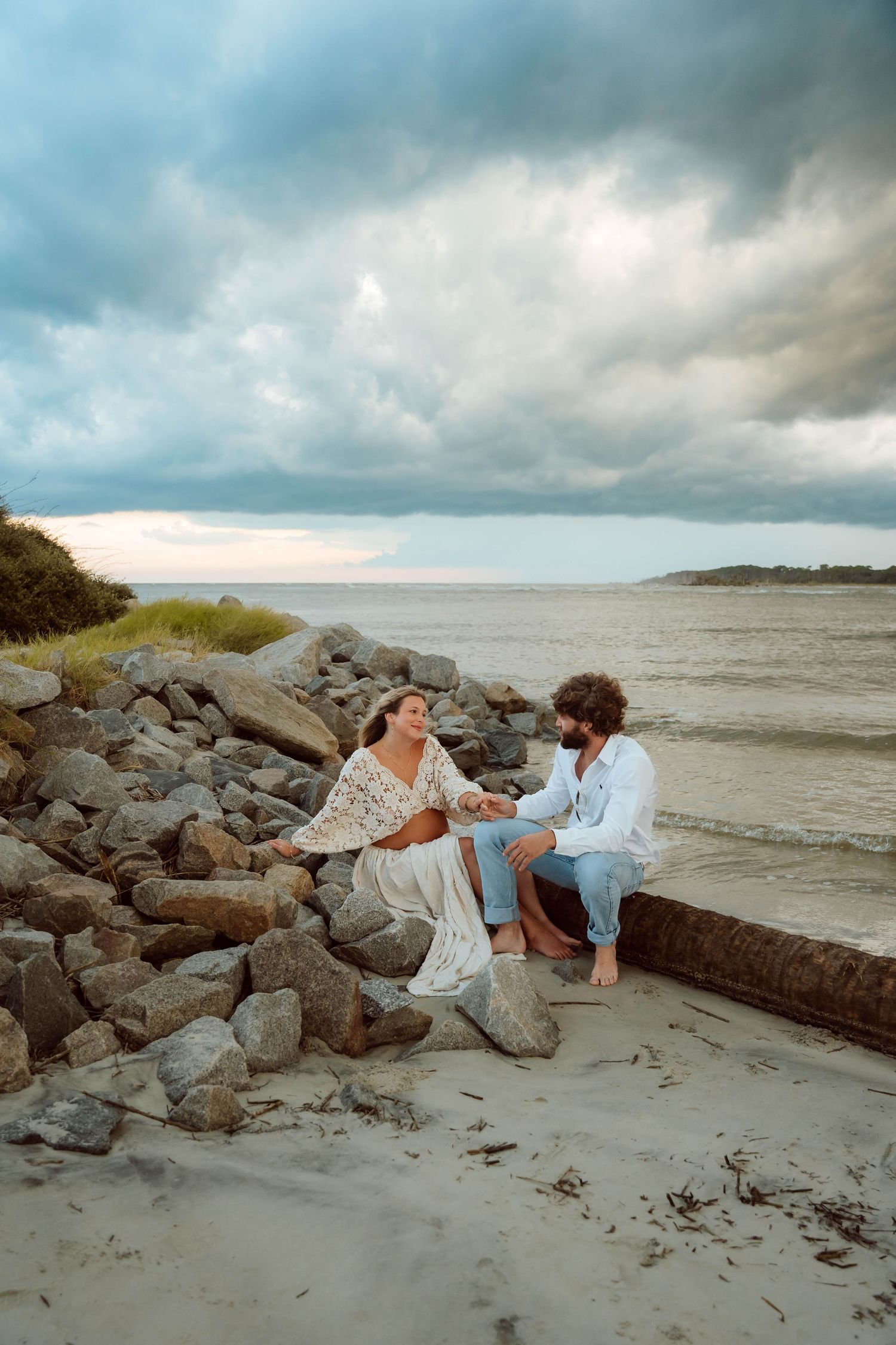 Couple relaxes on rocky beach coastline during moody cloudy afternoon.