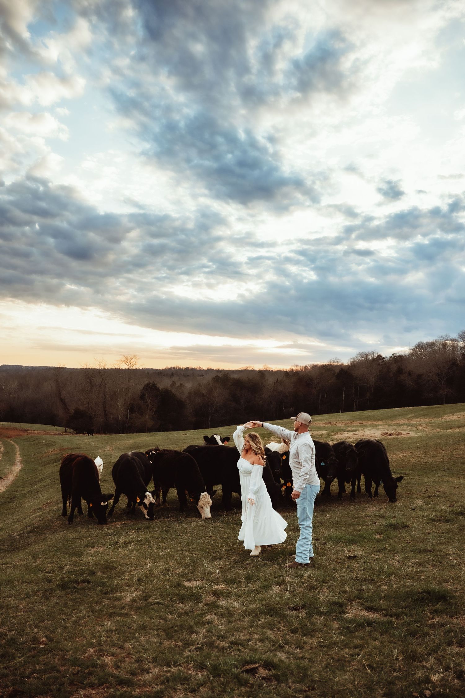 A couple in western attire poses with a herd of black cattle on a rural hillside at sunset.