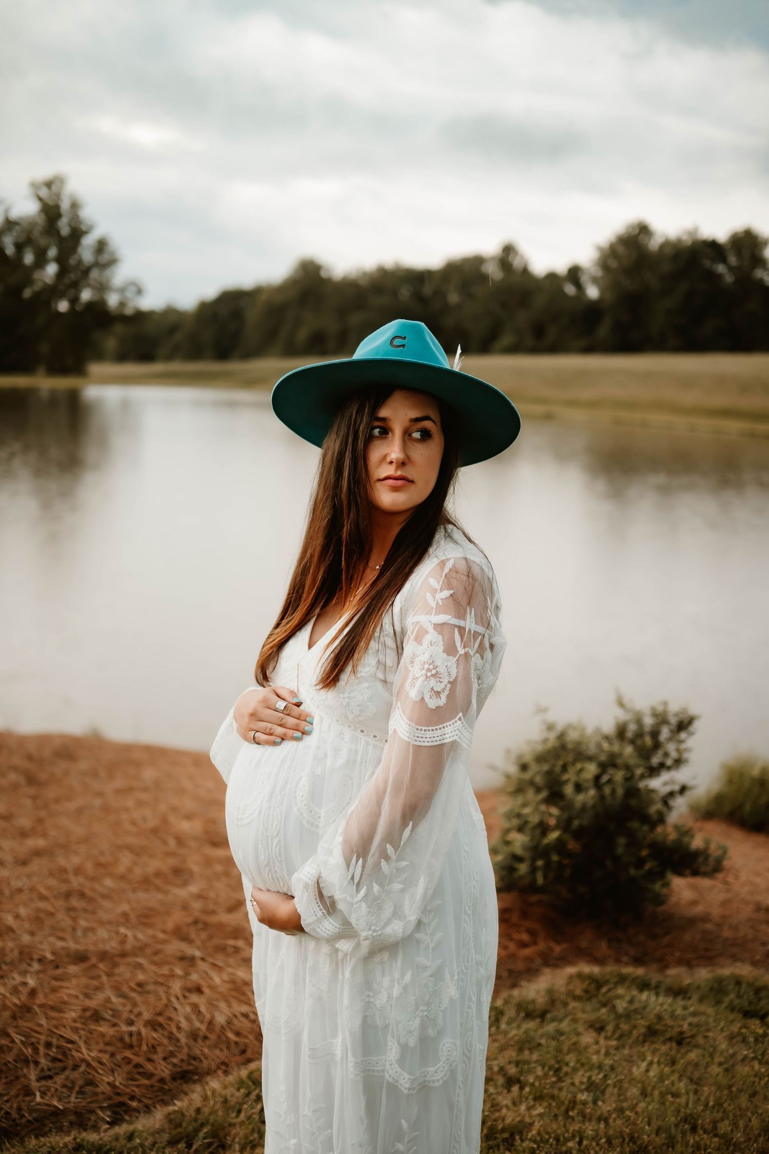 A moody maternity portrait by a lake featuring a white lace dress and wide-brimmed hat.