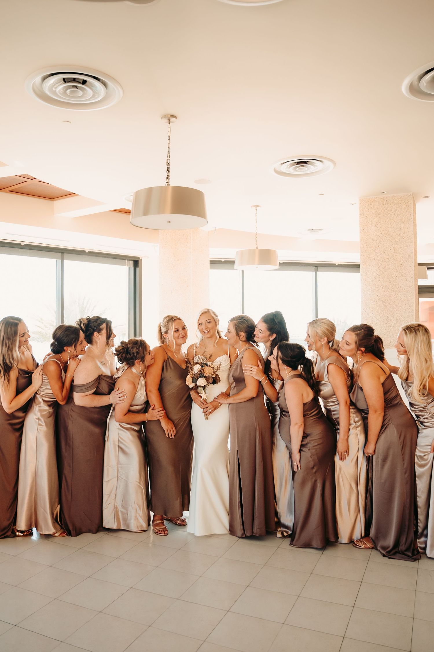 Wedding party in neutral brown dresses poses in line with backs to camera.