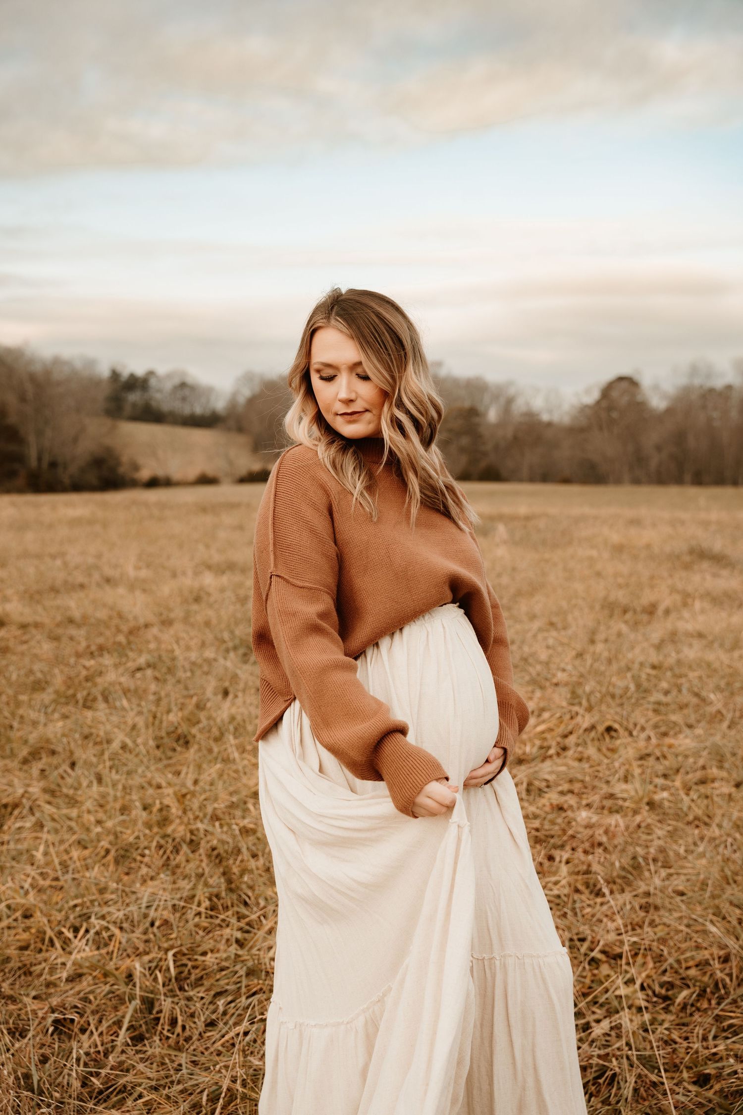 A woman in a brown sweater and white dress poses gracefully in a golden autumn field at sunset.