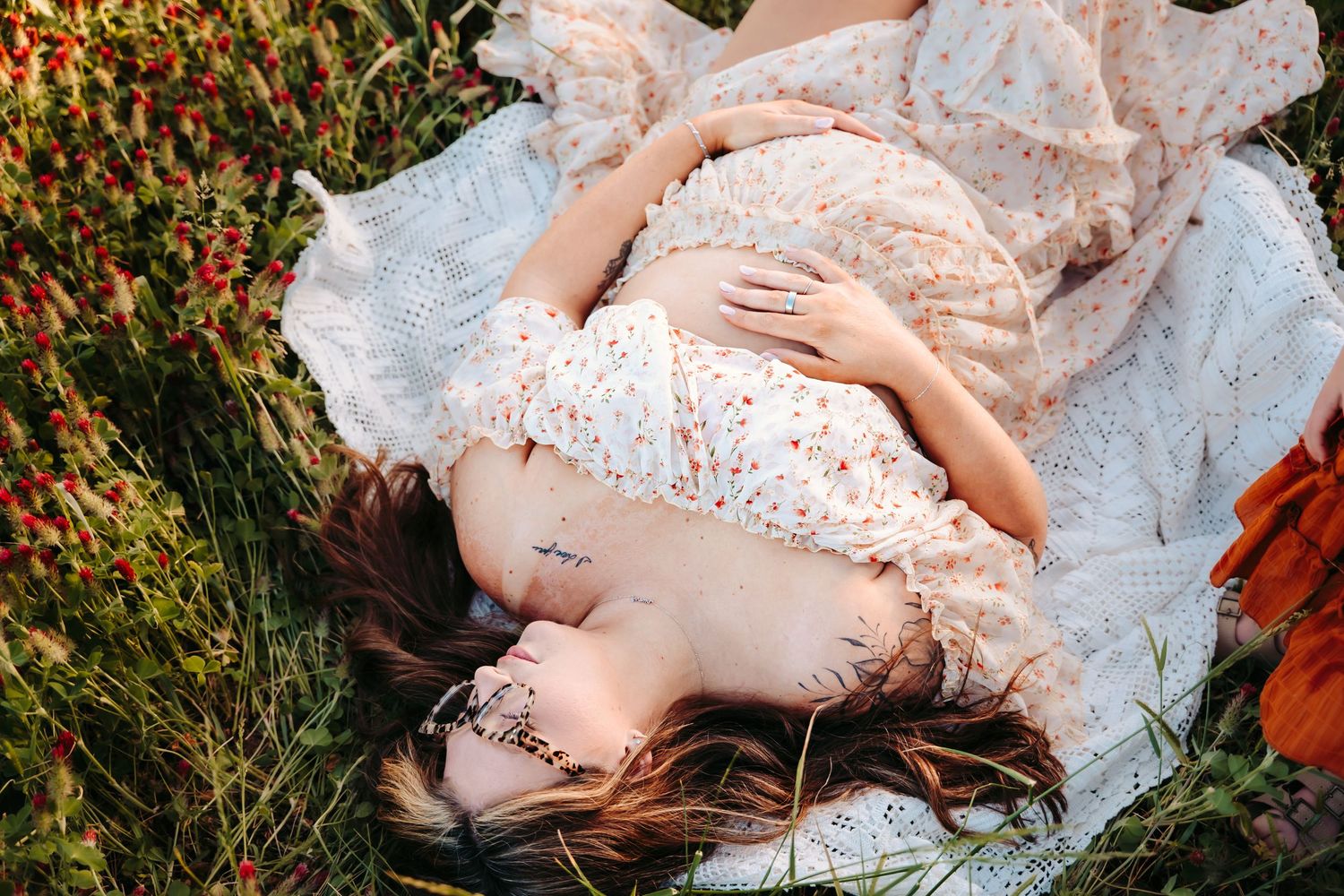 Pregnant woman in floral dress lying on white blanket in green field, cradling baby bump with hands.