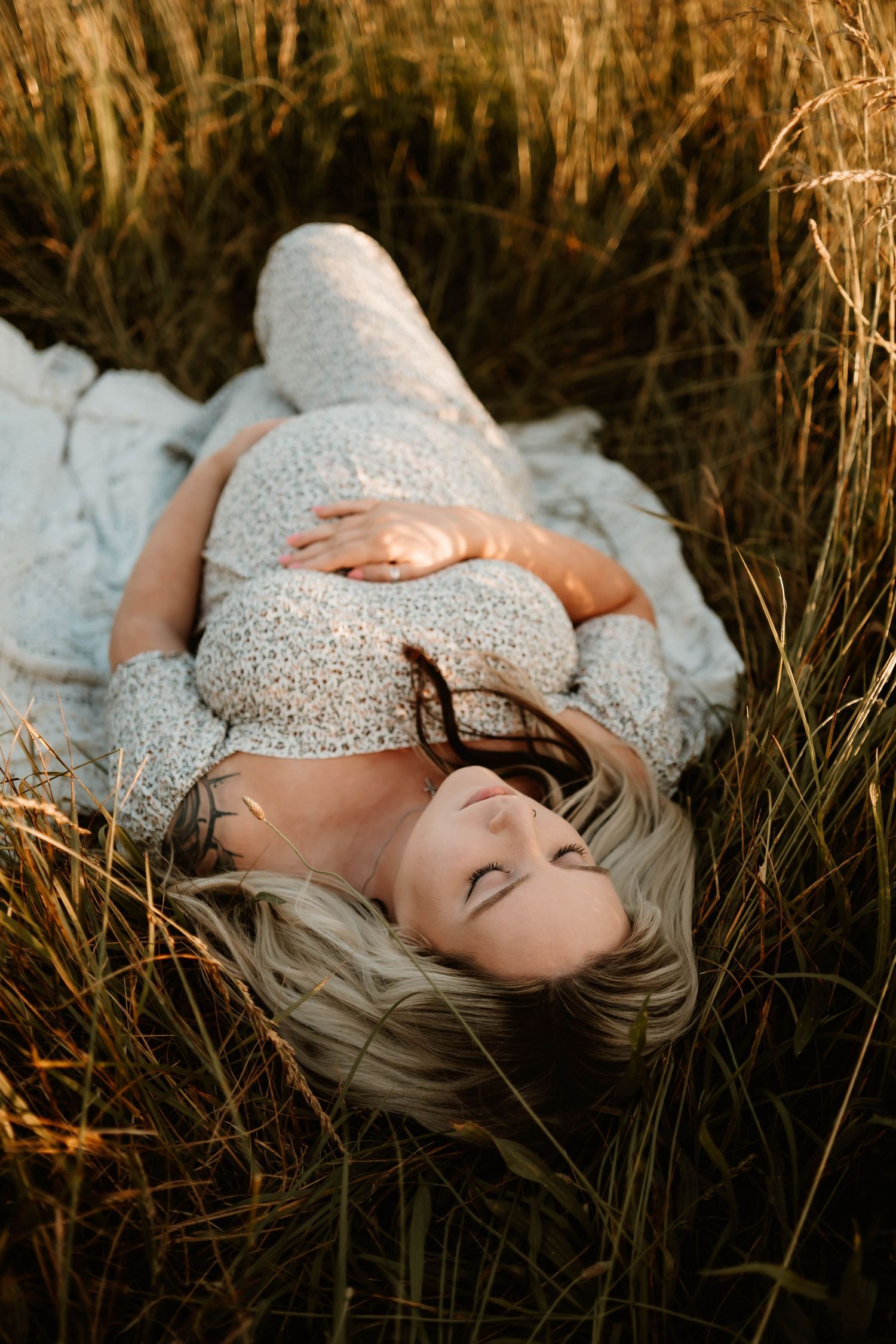 A dreamy maternity portrait of a woman in a white lace dress lying in tall grass at sunset.