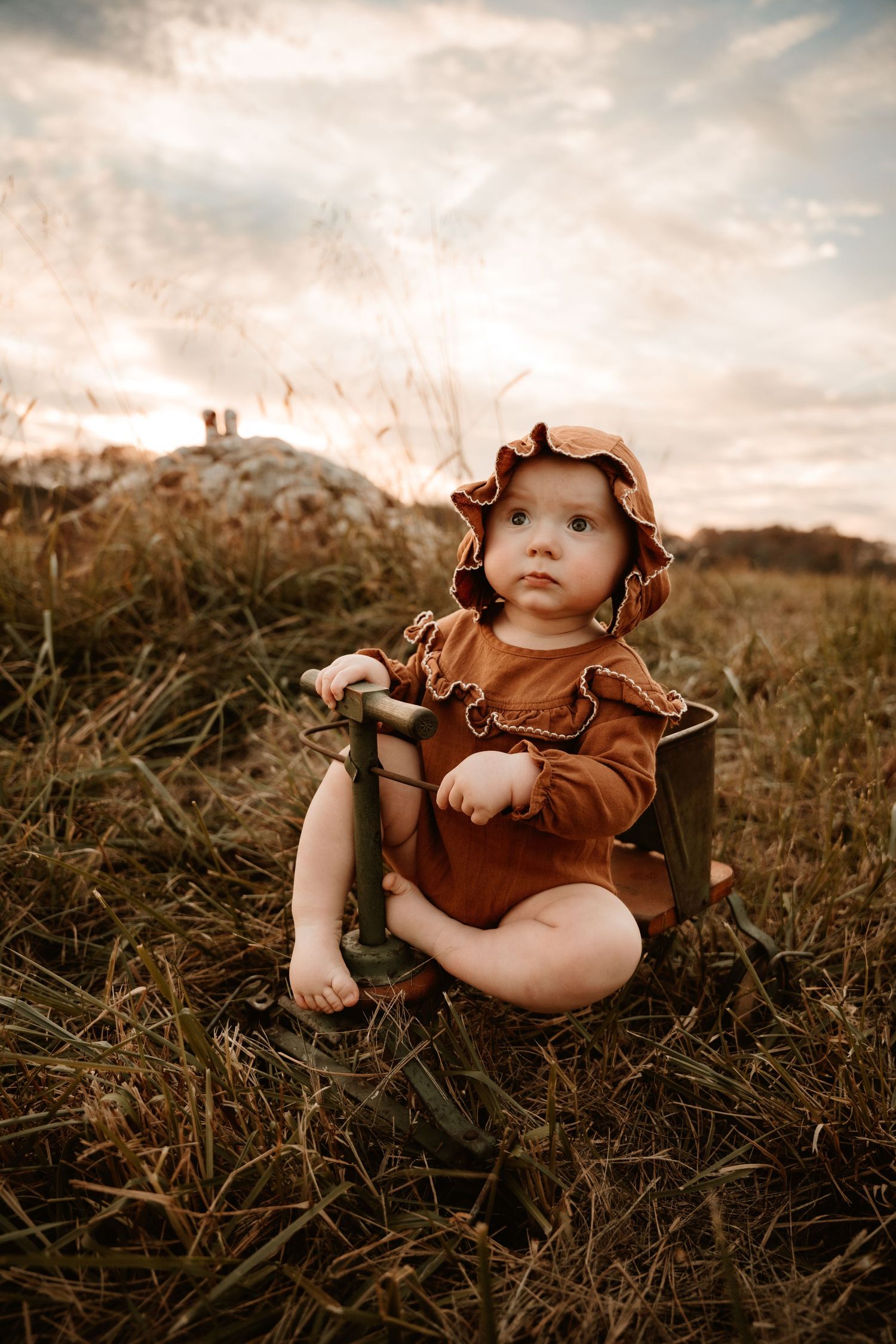 Baby in ruffled brown outfit playing in tall grass during sunset photoshoot.