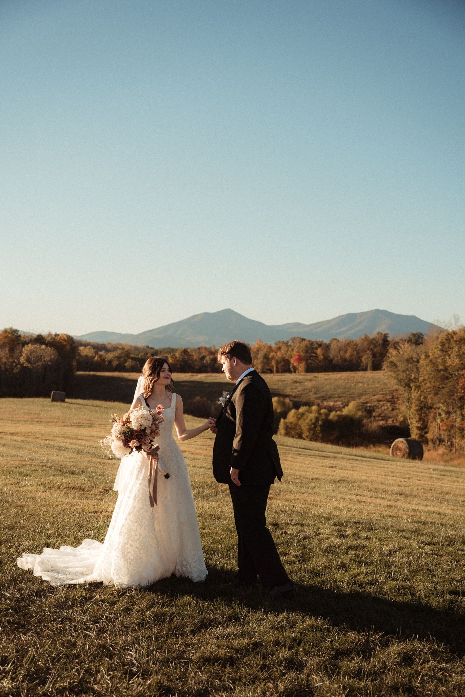 Couple in wedding attire standing in mountain meadow during autumn golden hour.