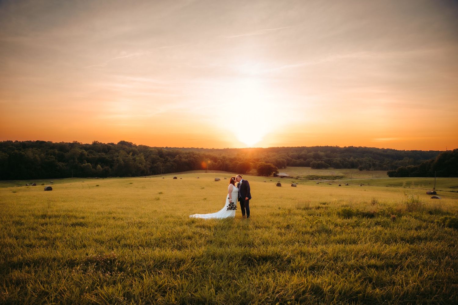 Silhouette of newlyweds standing in a meadow during a dramatic sunset with rolling hills in the background.
