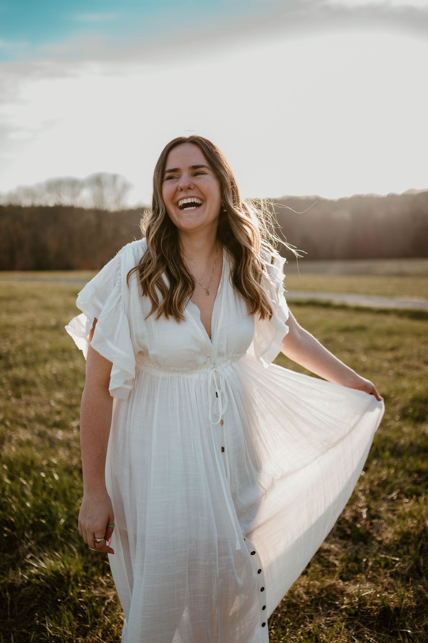 Person in flowing white dress laughing and twirling in a sunlit meadow at golden hour.