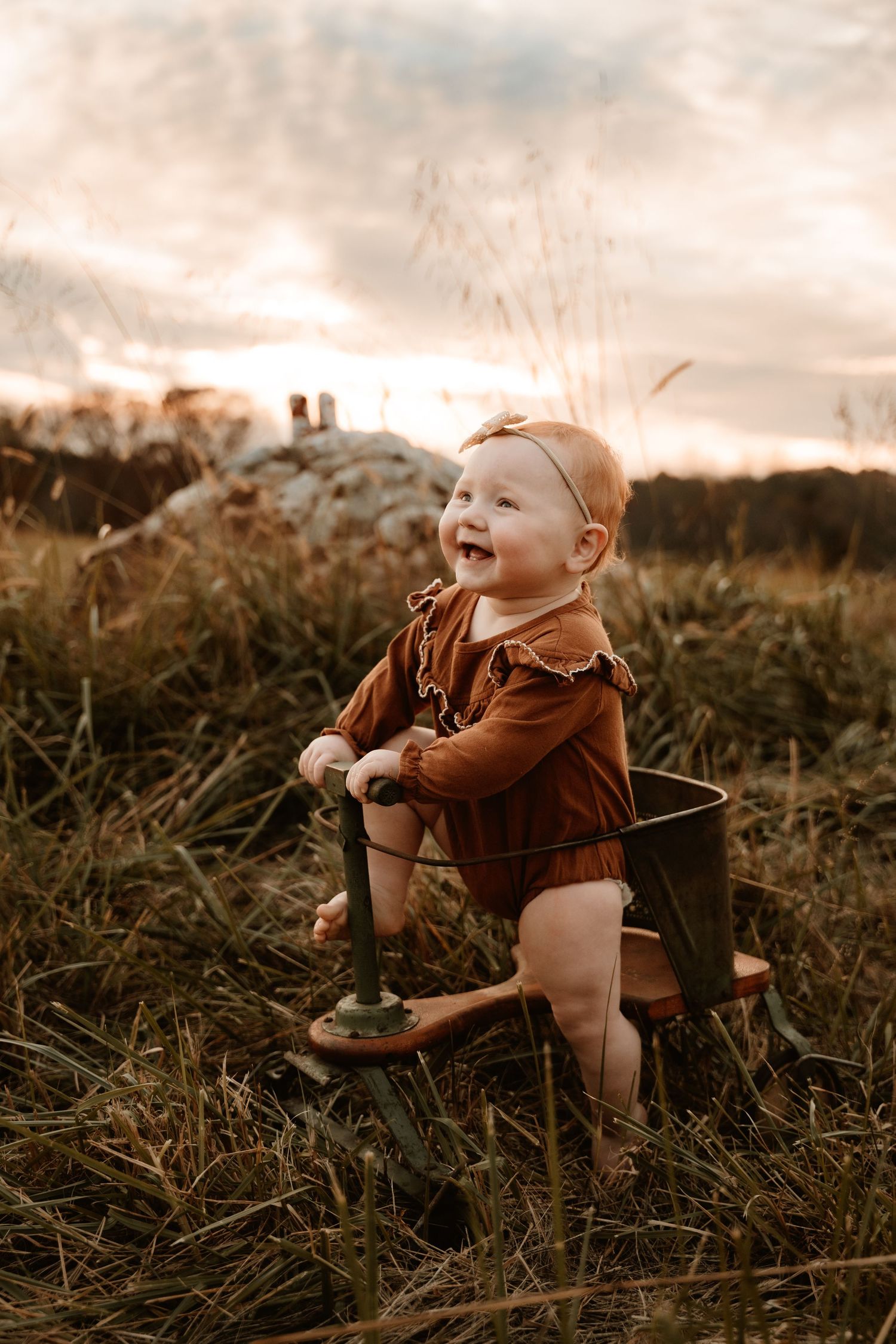 Baby in brown outfit sitting and laughing in tall grass during sunset.