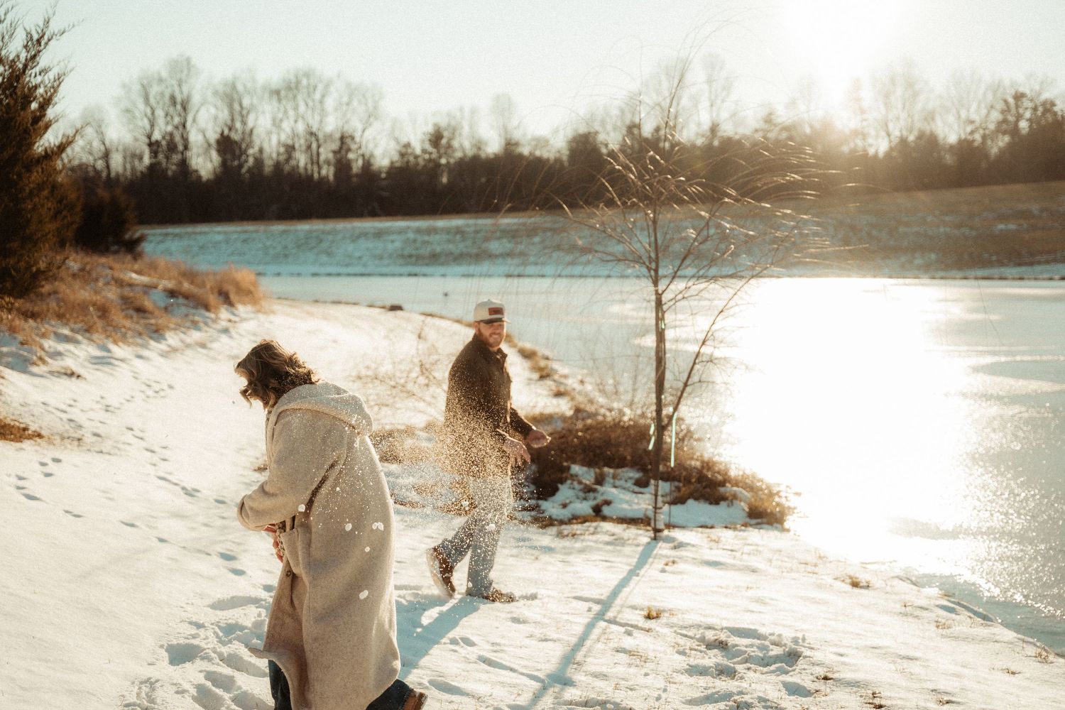 Two people walk along a snowy riverbank on a bright winter day.