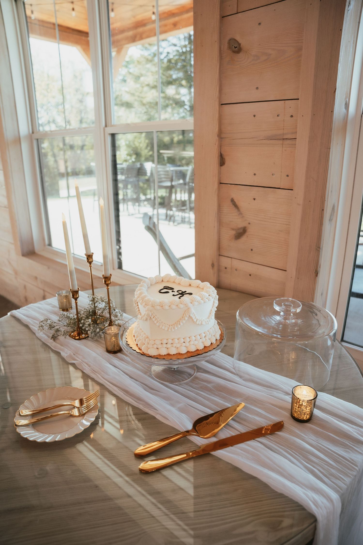Simple white wedding cake on a rustic wooden table with gold utensils and natural window lighting.