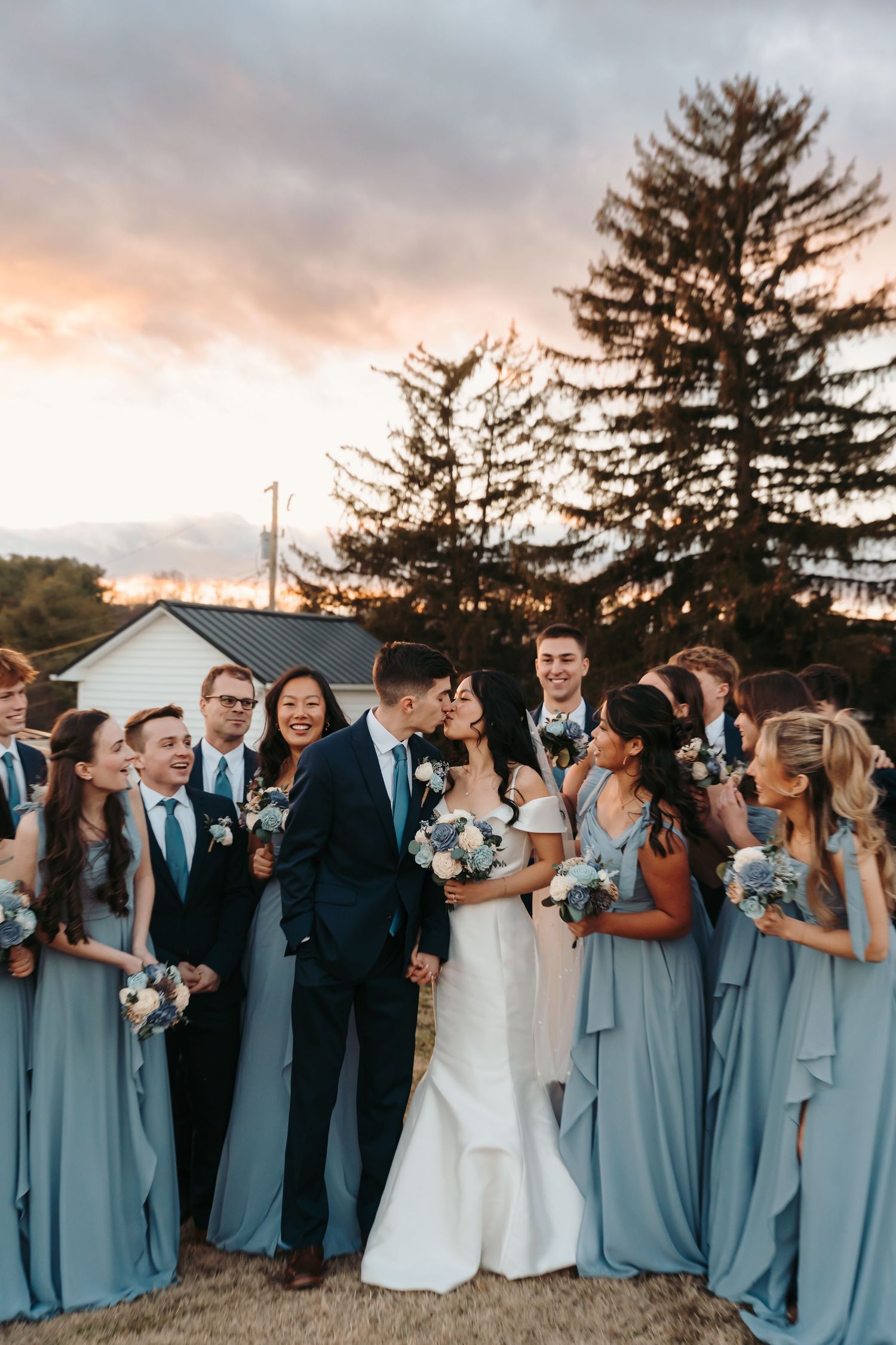 Wedding party in dusty blue bridesmaid dresses and black suits pose together against sunset sky.