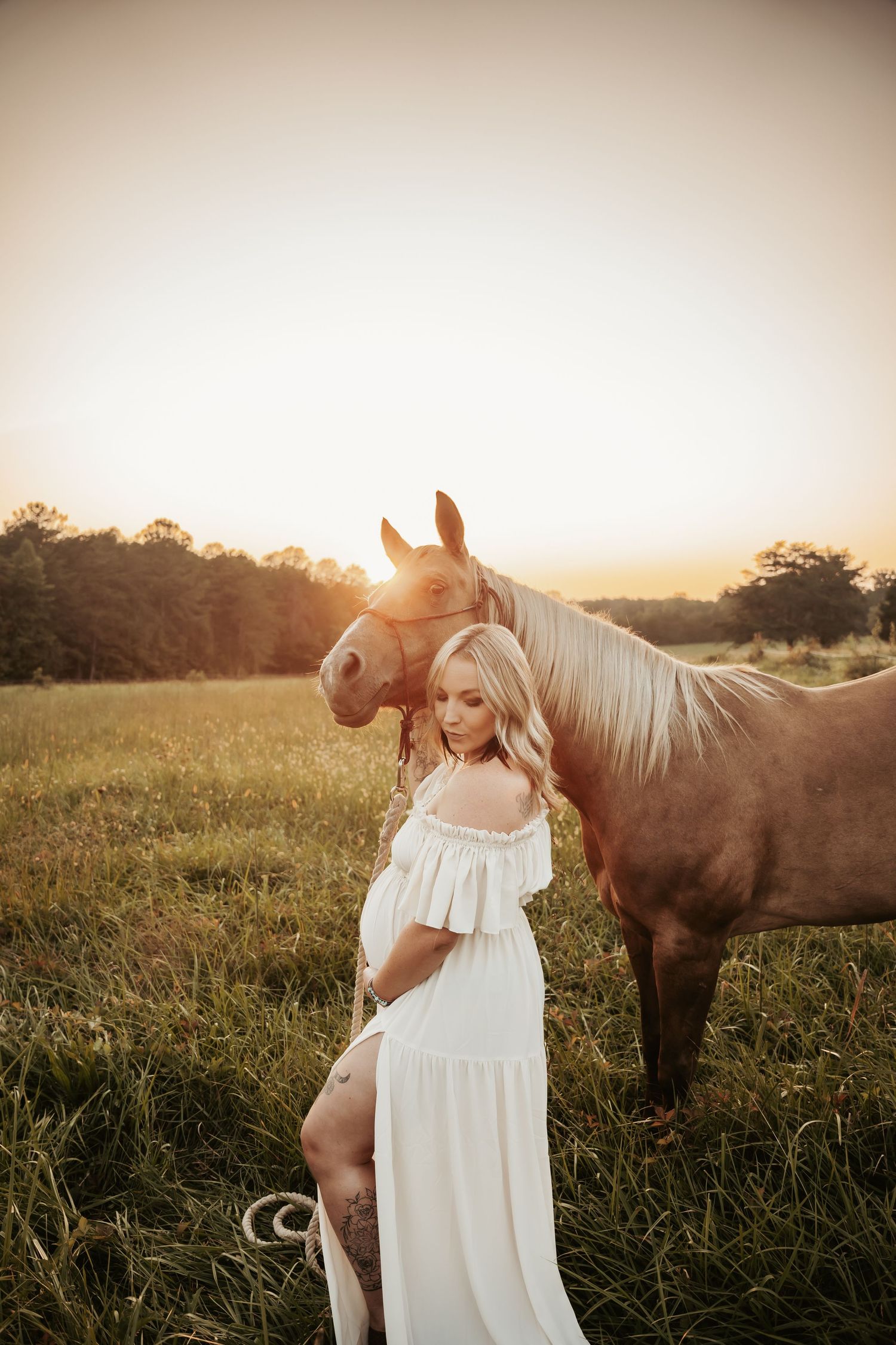 Woman in flowing white dress poses with brown horse in sunlit field at dusk.