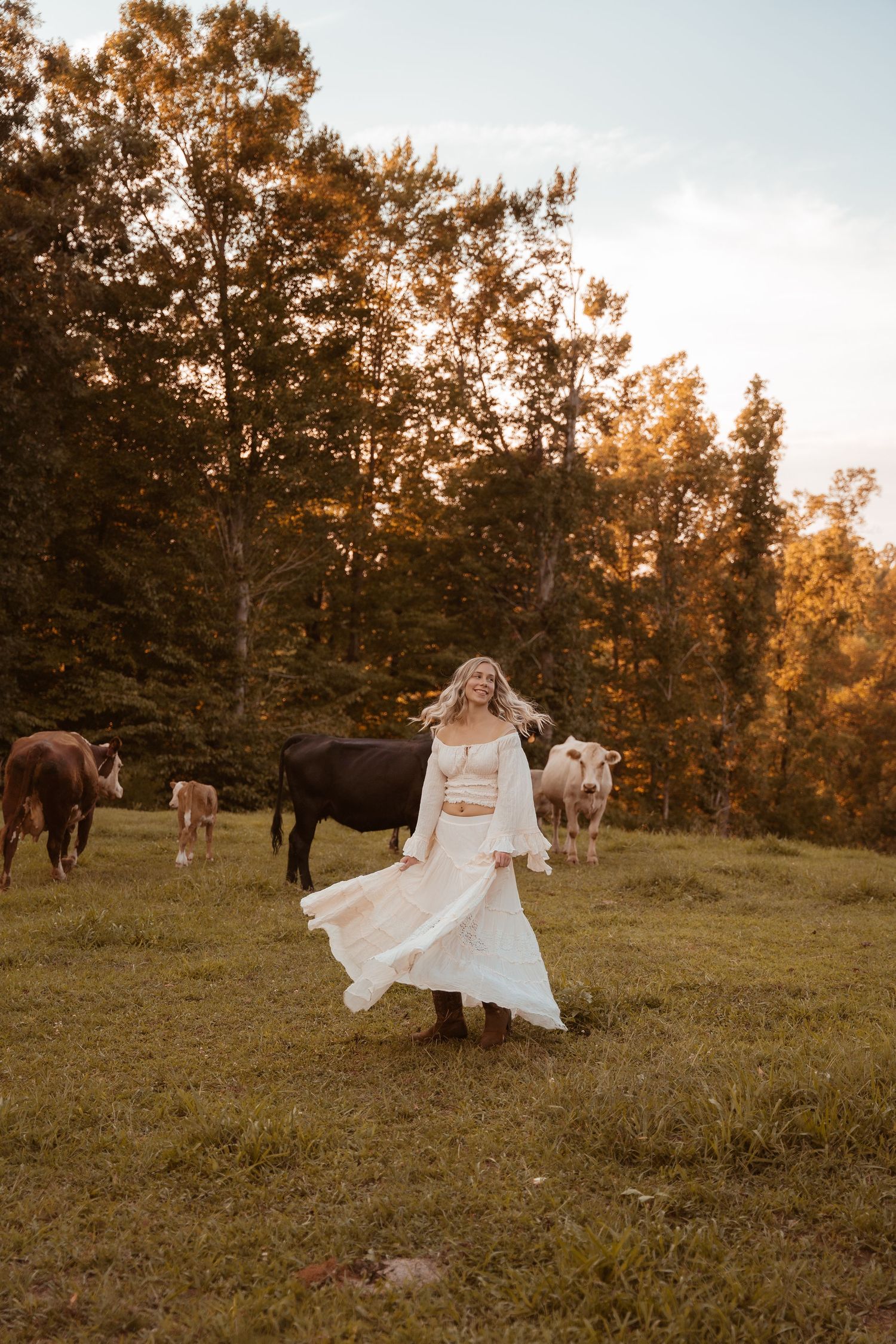 White dress twirls among grazing cows in autumn field at sunset.