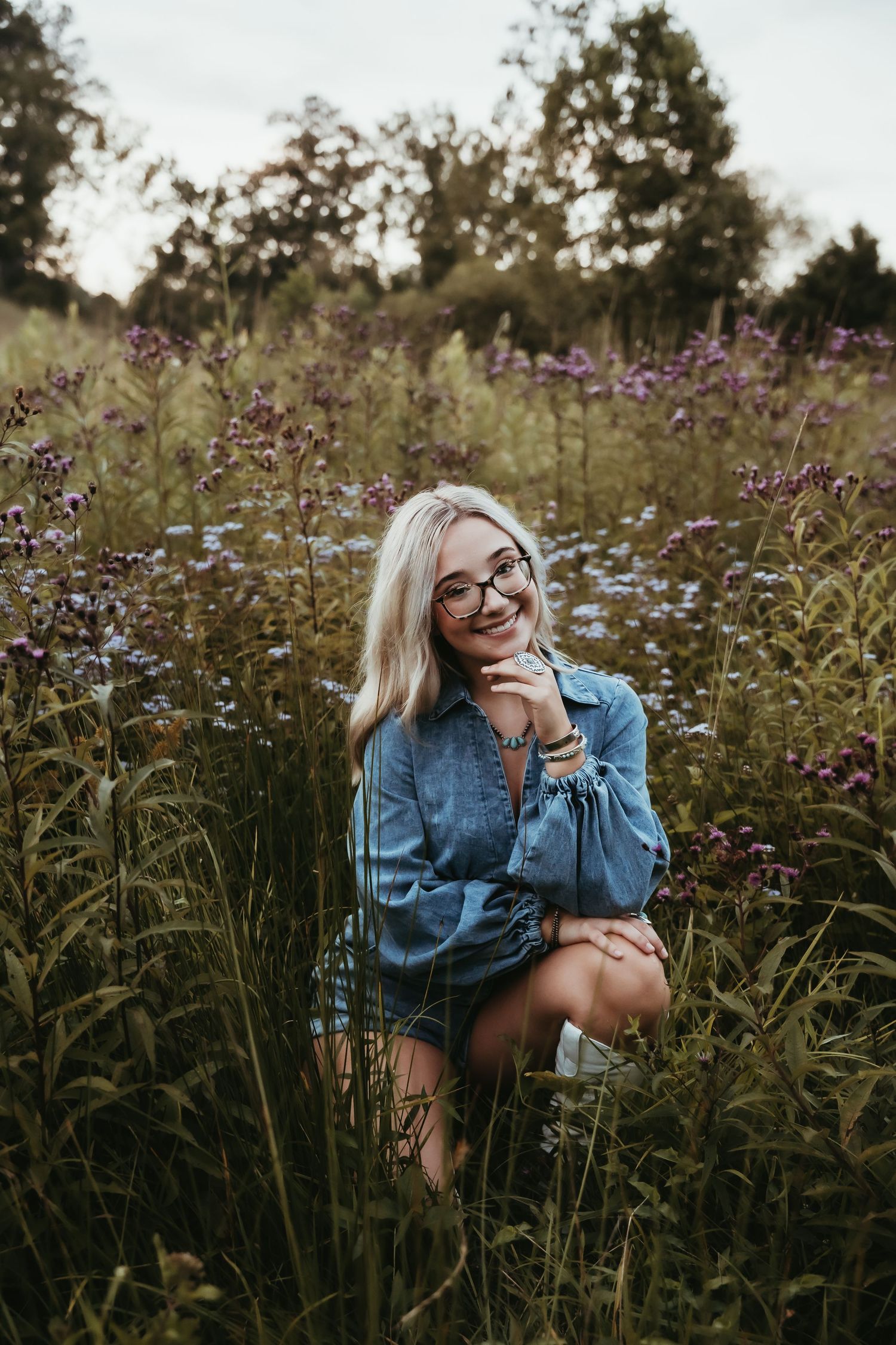 Smiling blonde woman wearing glasses and denim shirt sitting in a wildflower field with purple blooms.