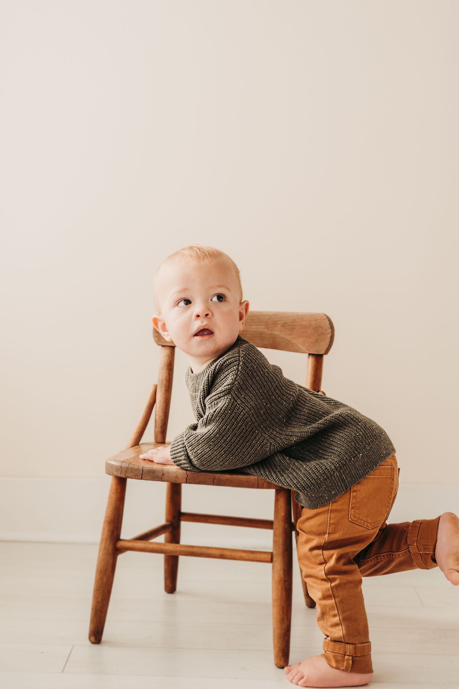 Small child in gray sweater poses playfully on wooden chair against white background.