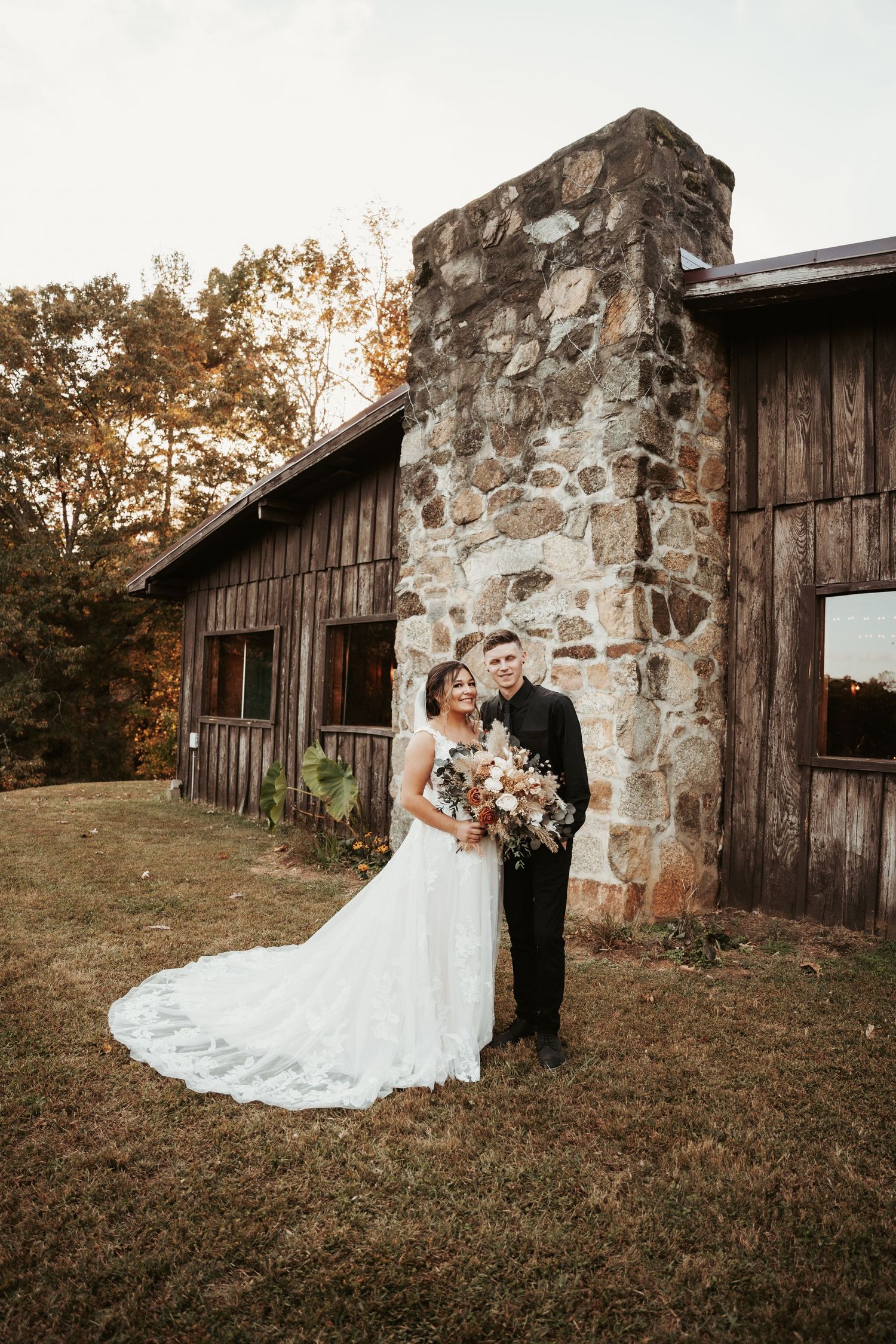 Wedding couple poses in front of rustic stone and wooden barn wall during autumn photoshoot.