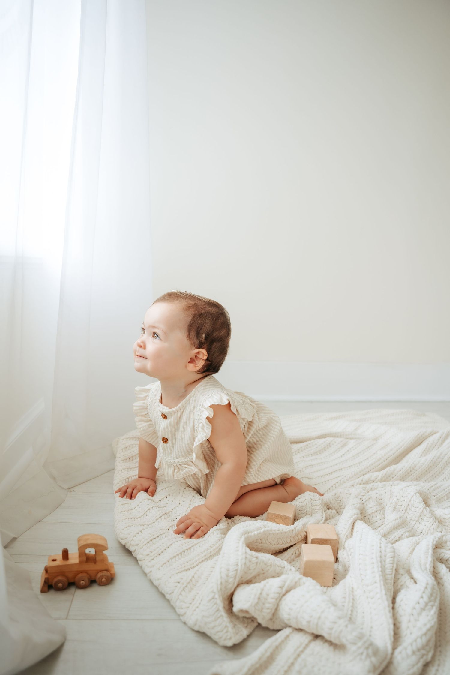 Baby in white outfit lying on textured bedding near window with soft natural light.