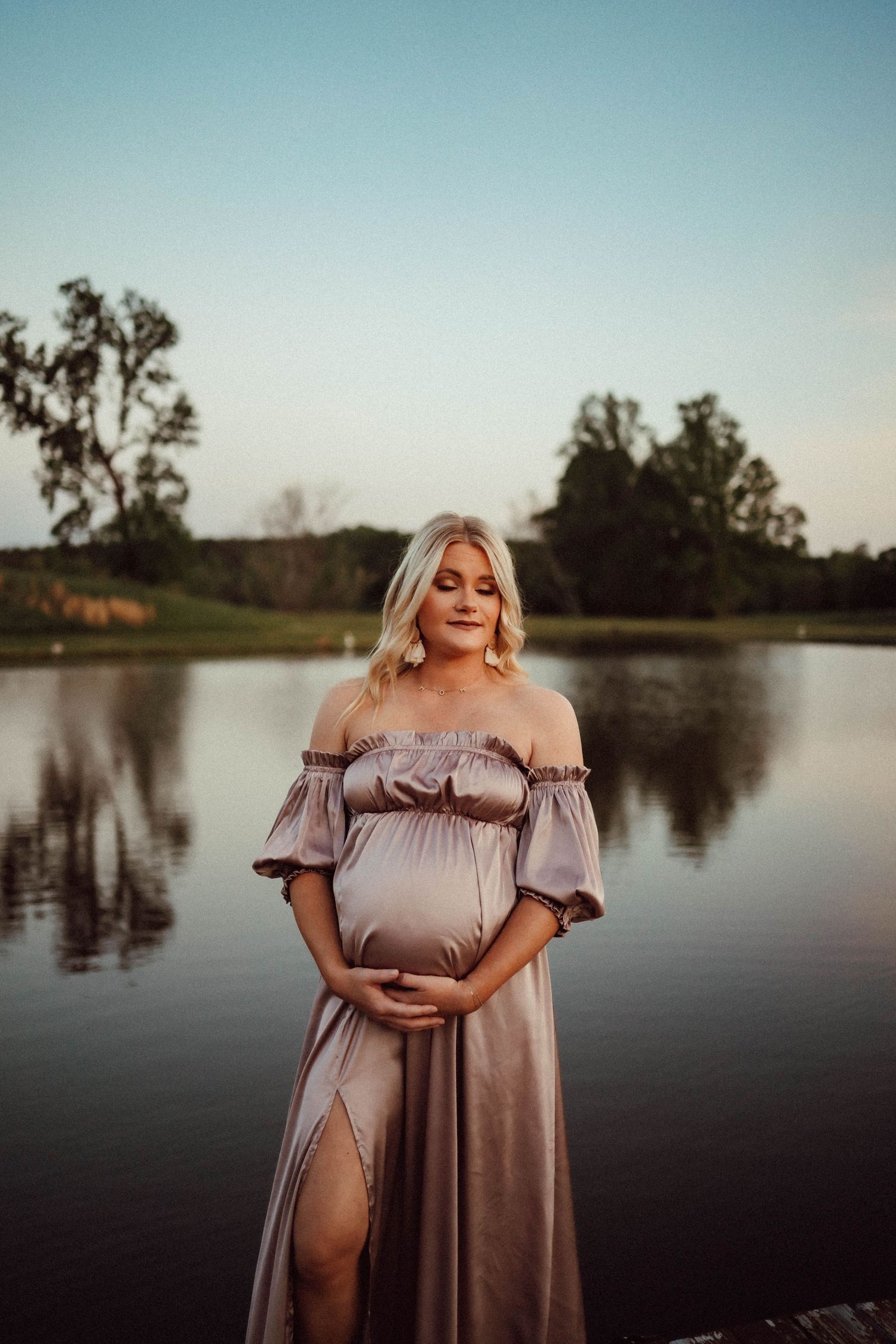 Maternity photoshoot at sunset by a serene lake featuring a flowing mauve dress.